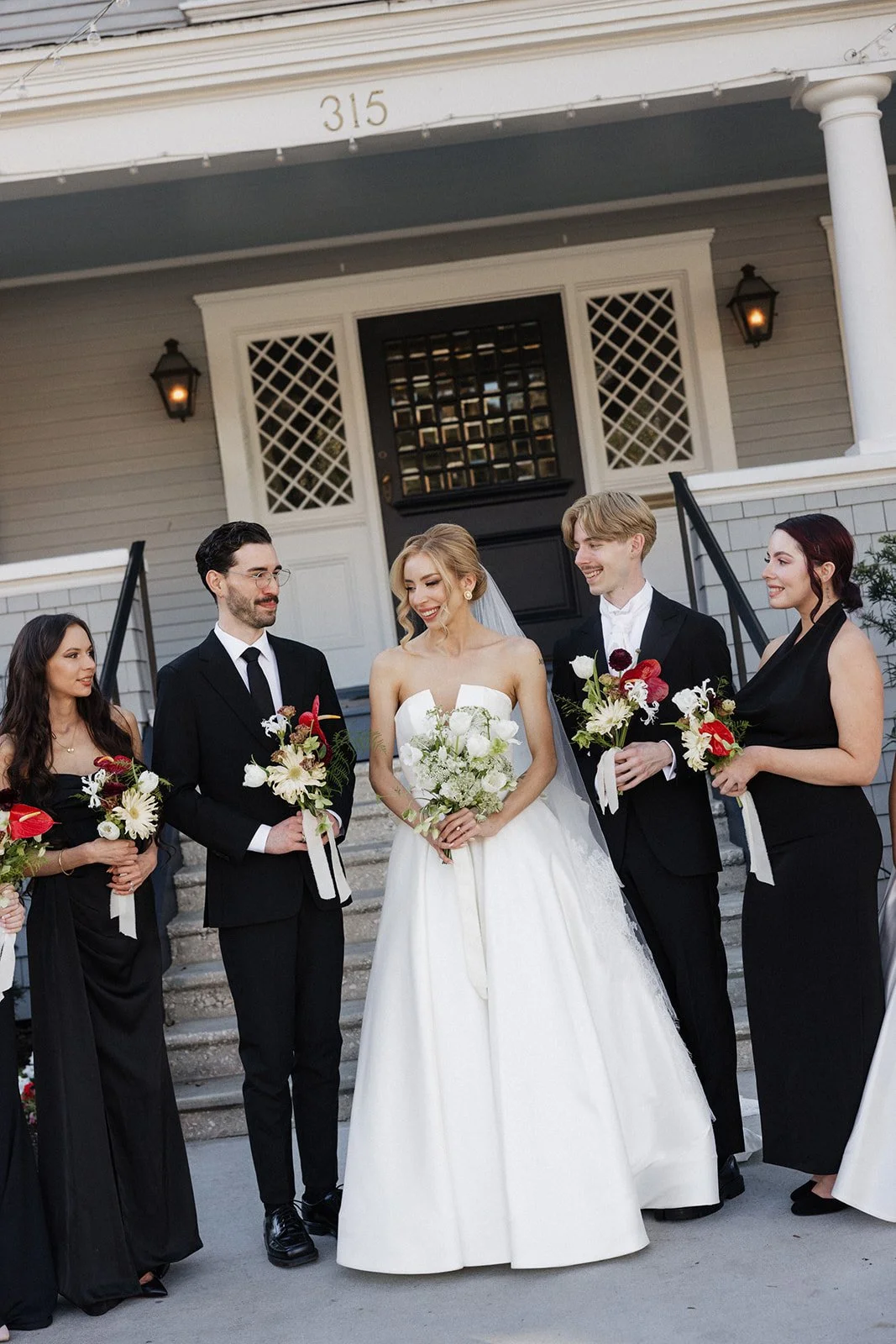 A wedding group outside a house, with the bride in a white dress holding a bouquet, surrounded by bridesmaids and groomsmen in formal black attire holding small bouquets, standing on stairs in front of a gray house with a black door.