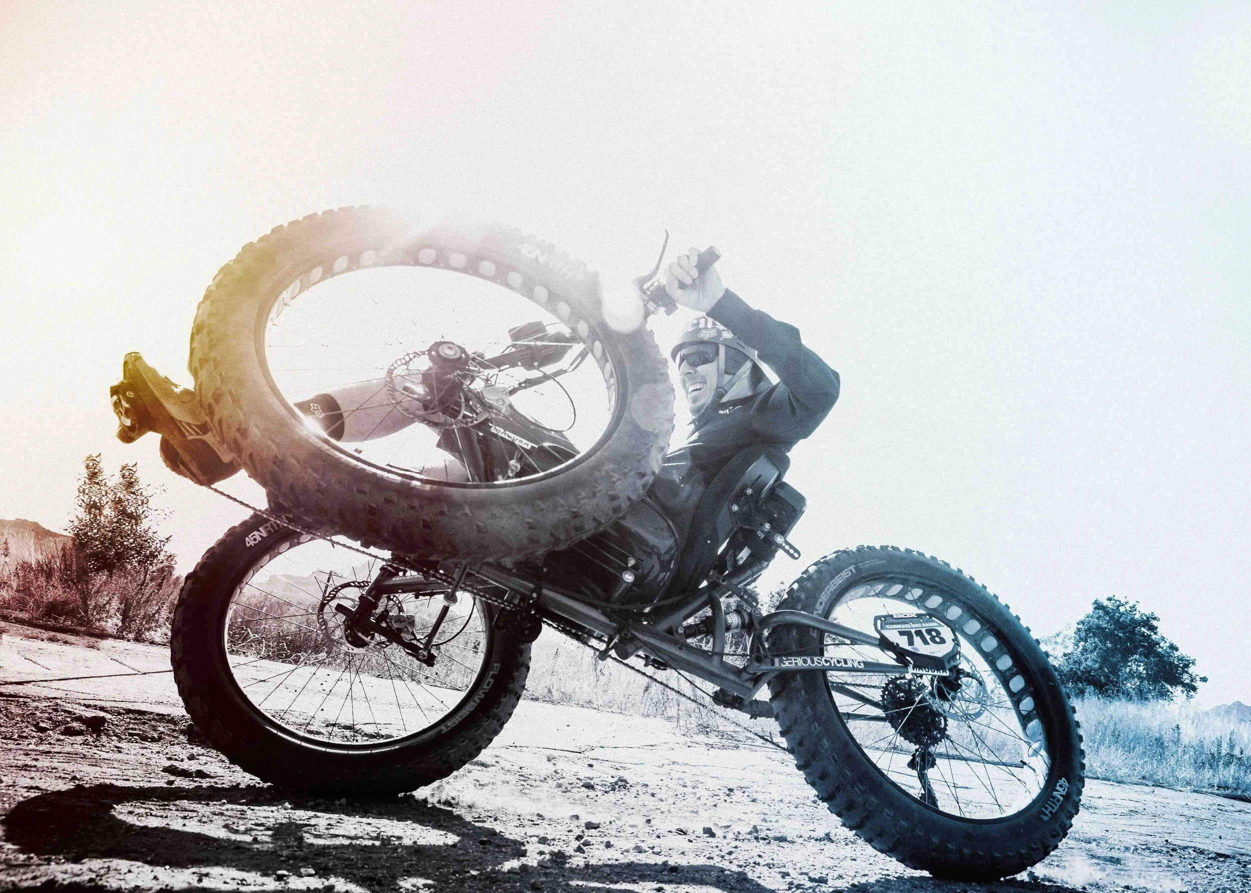 Aaron Baker, a mountain biker in black gear and helmet performing a wheelie on a dirt trail in a desert landscape.