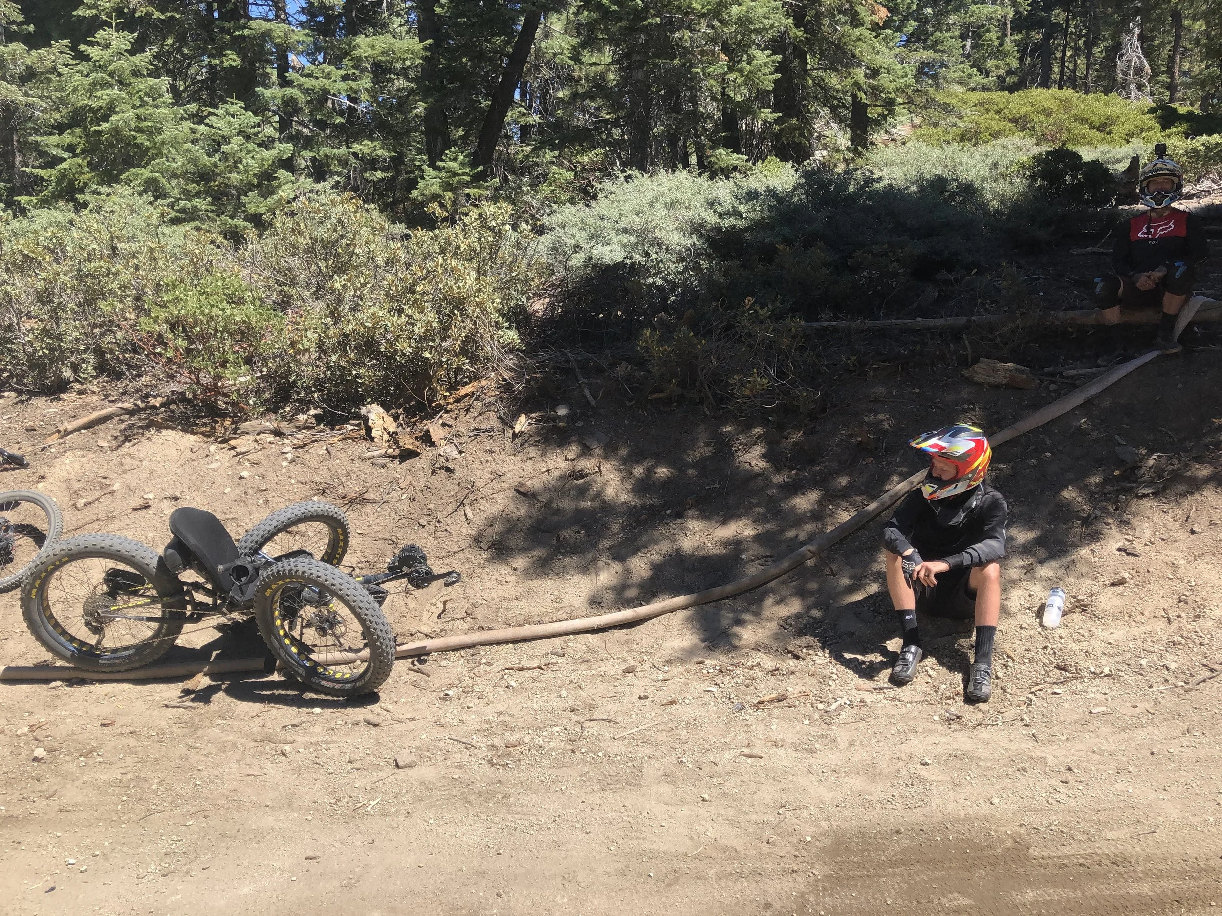 A trail in a forested area with two mountain bikes fallen on the ground, and a person sitting on the trail wearing a helmet, black gear, and a helmet with goggles, surrounded by trees and bushes.