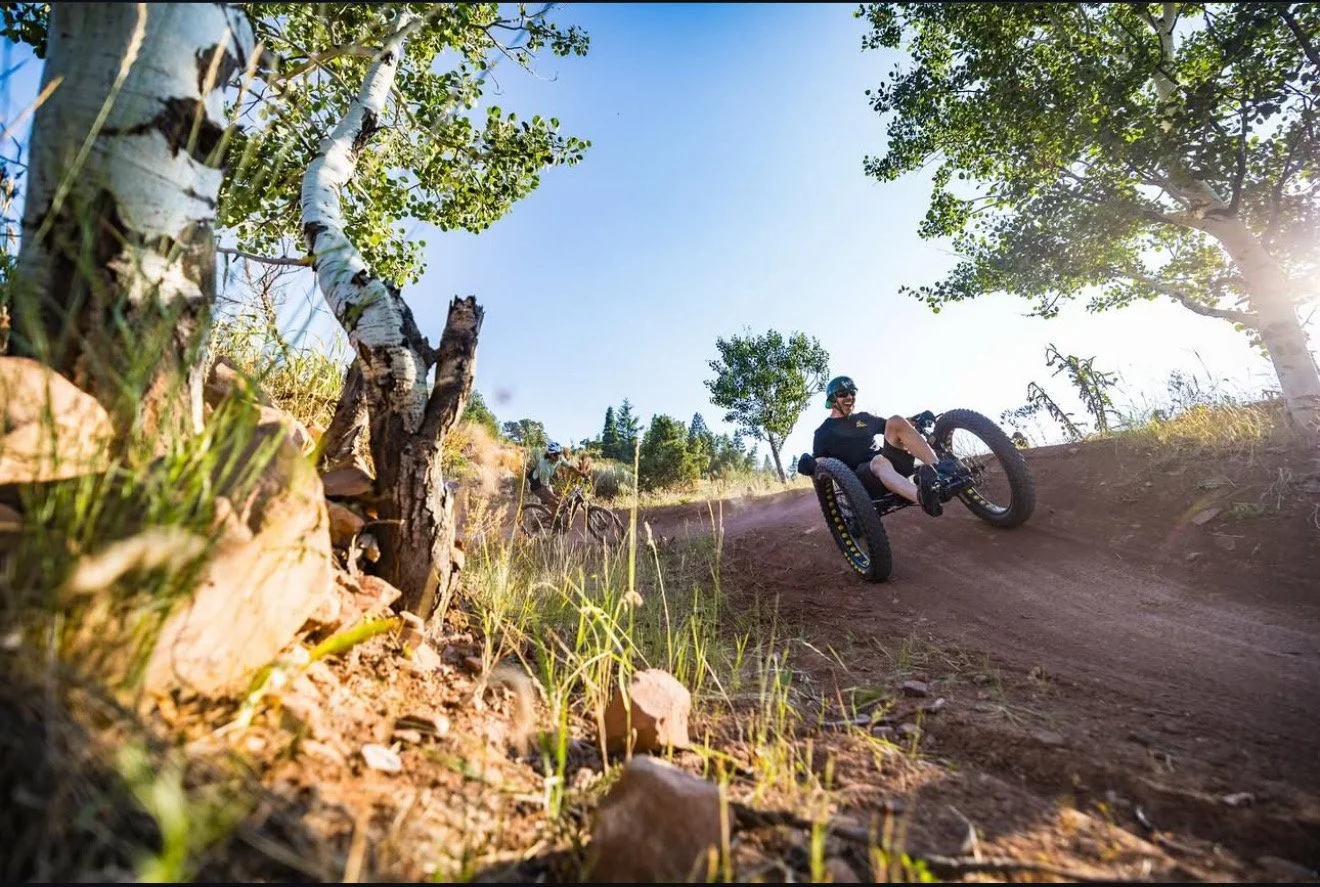 Aaron Baker and two mountain bikers riding on a dirt trail surrounded by trees and rocks on a sunny day.
