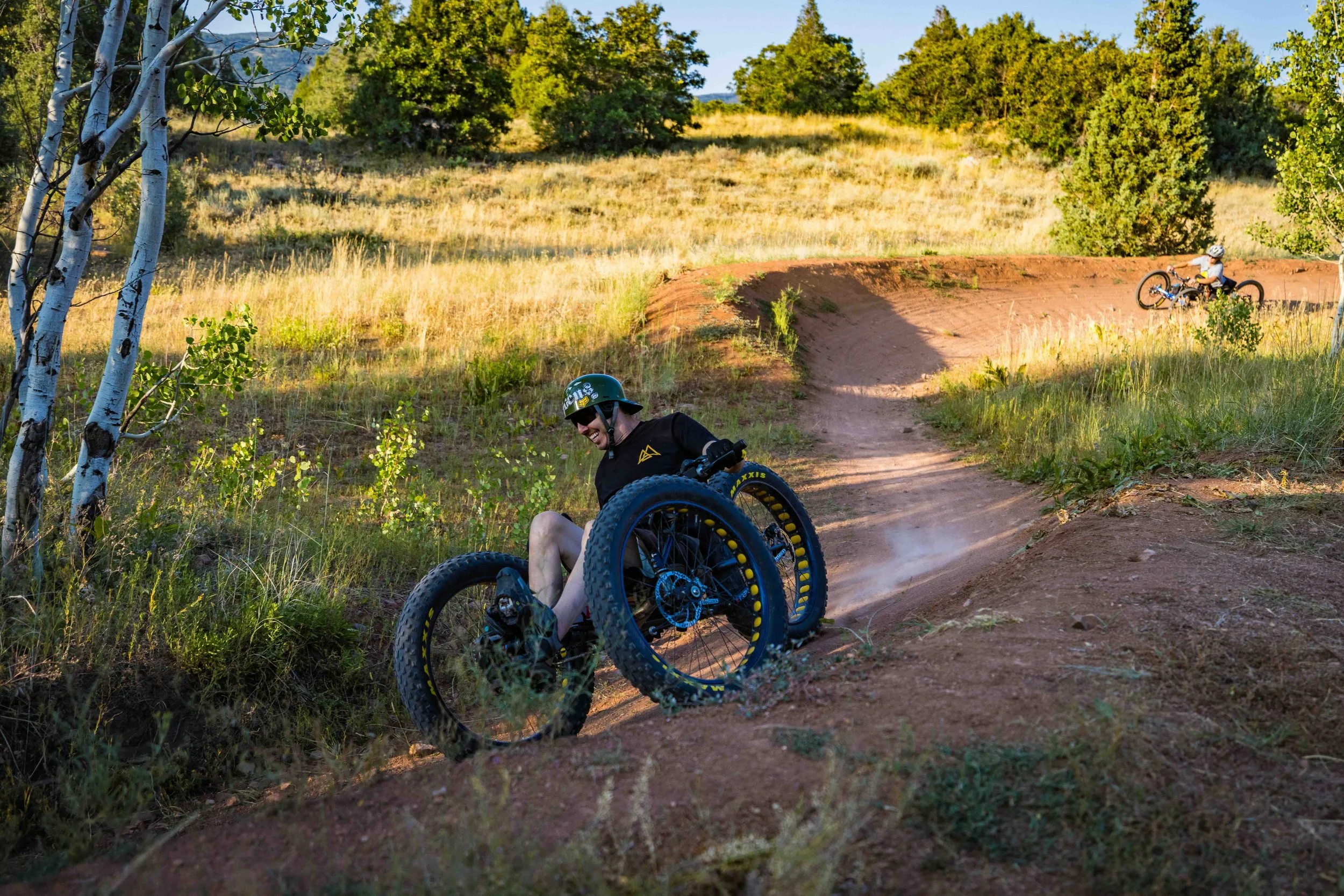 Aaron Baker in a black shirt and helmet falling off a mountain bike on a dirt trail during daytime, with another person riding a bike in the background, in a natural landscape with trees and grassy fields.