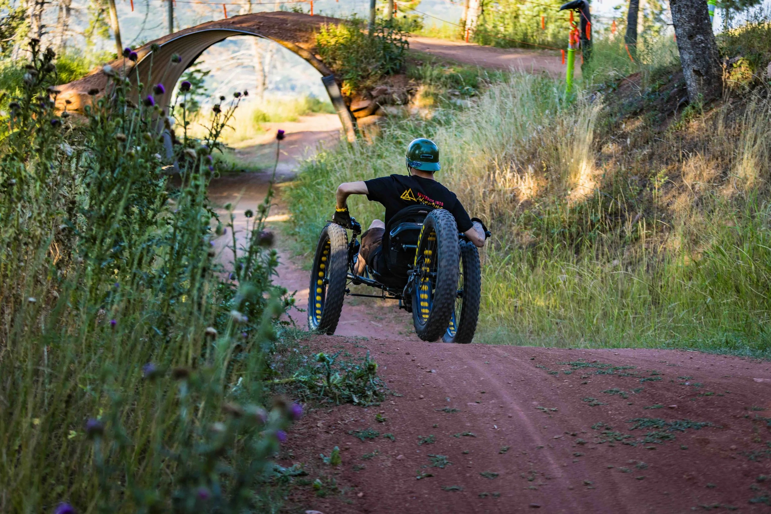 Aaron Baker riding on a dirt trail through a wooded area, passing under a small bridge with flowers and trees around.