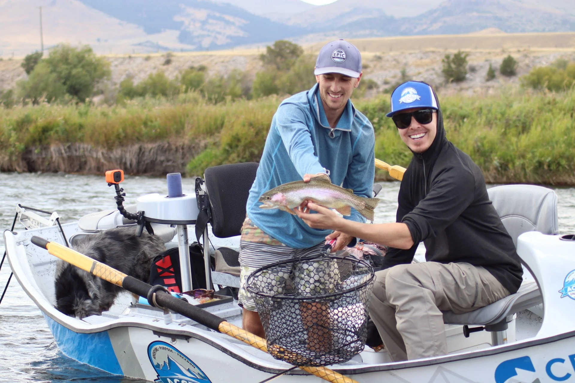 Two men on a boat holding a large fish, with a dog nearby, on a river with grassy banks and mountains in the background.