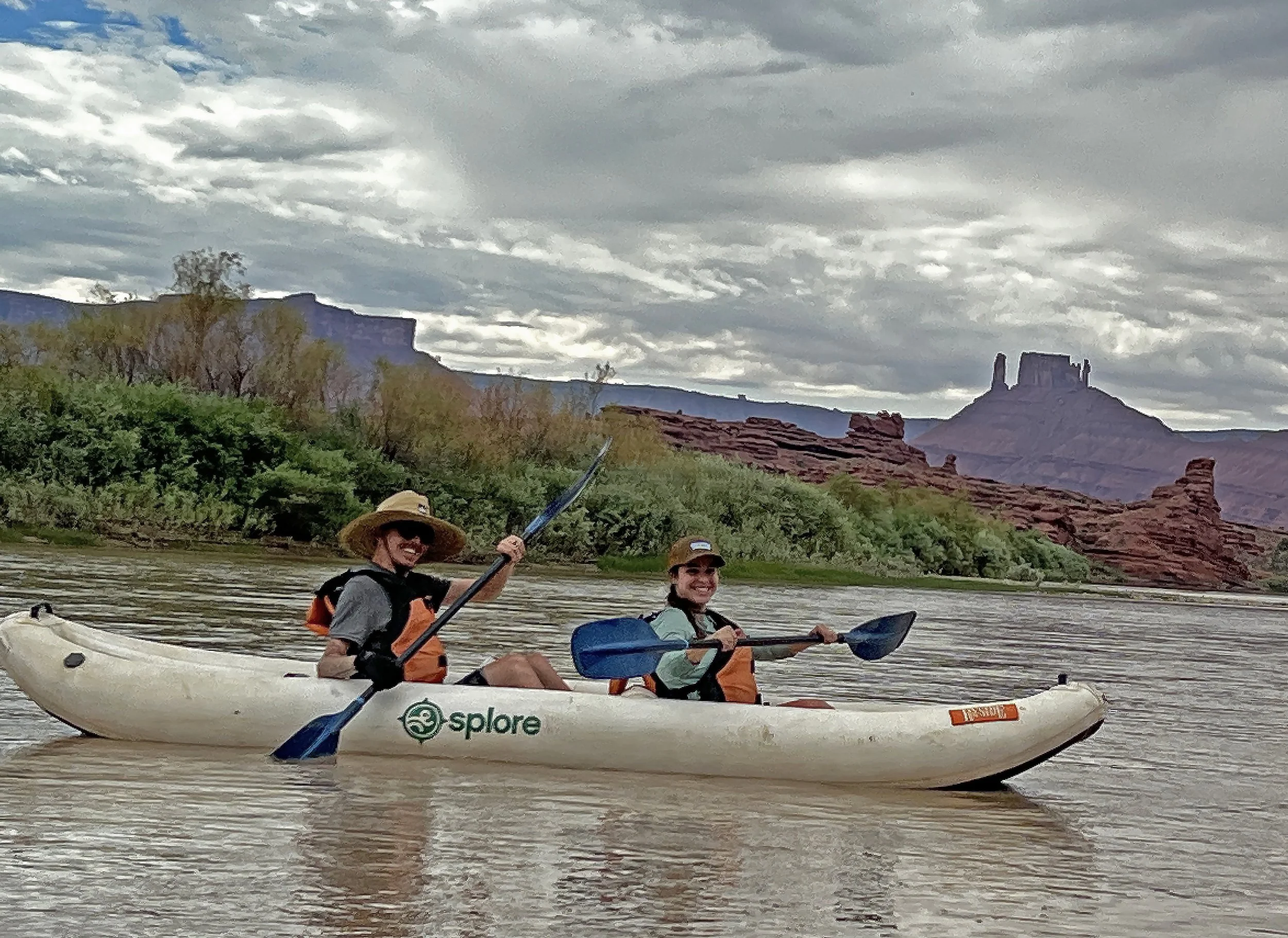 Aaron Baker kayaking on a river under a cloudy sky, with a scenic backdrop of red rock formations and distant cliffs.