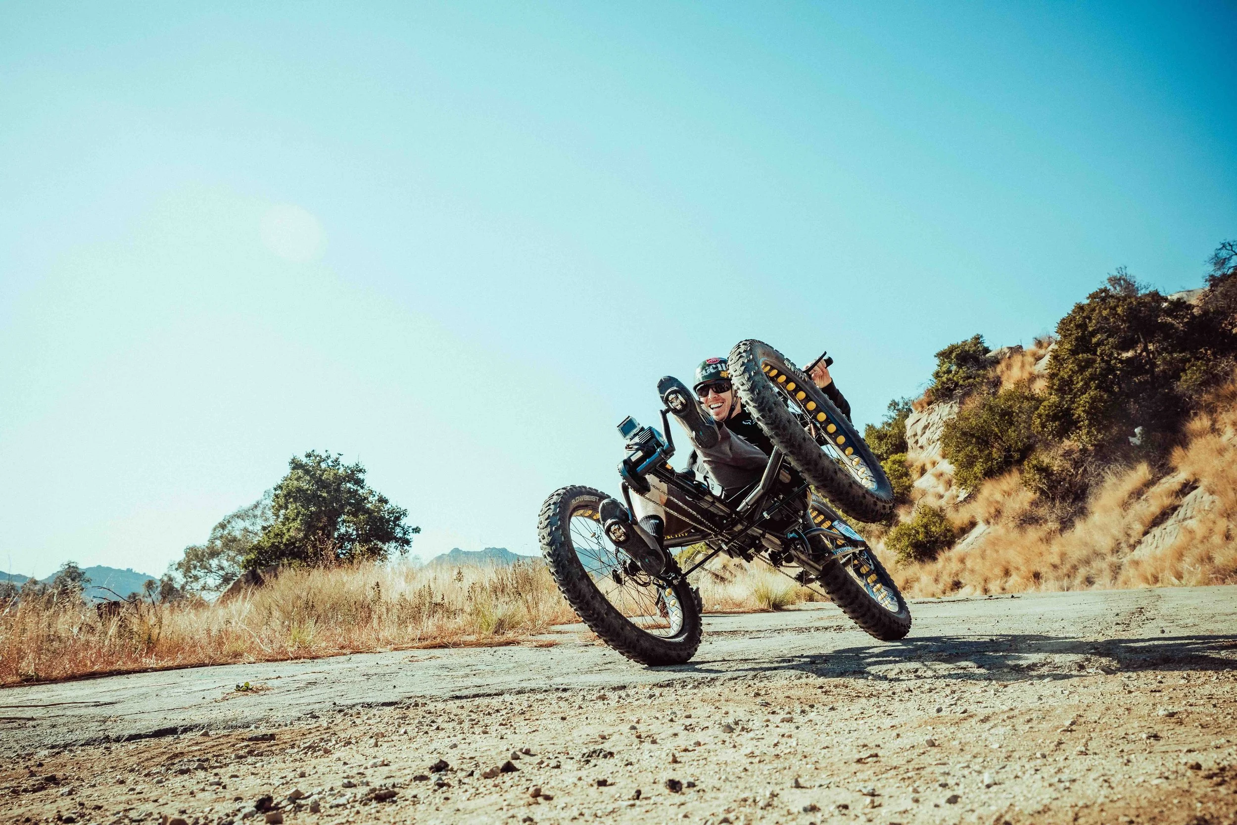 Aaron Baker performing a wheelie stunt on a mountain trail with trees and hills in the background.