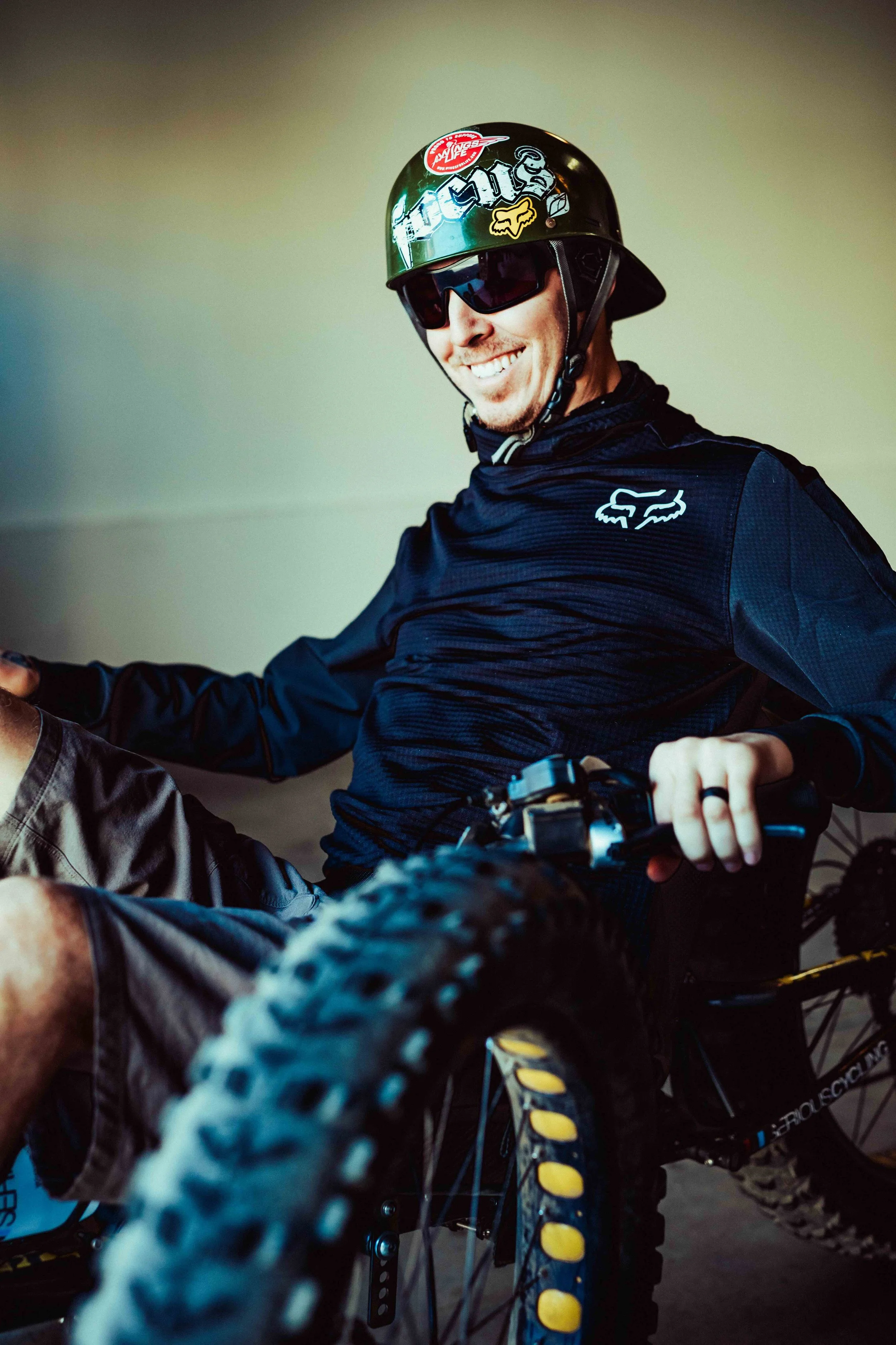 Aaron Baker wearing a black helmet with stickers, black sunglasses, and a black jacket, sitting on a mountain bike.
