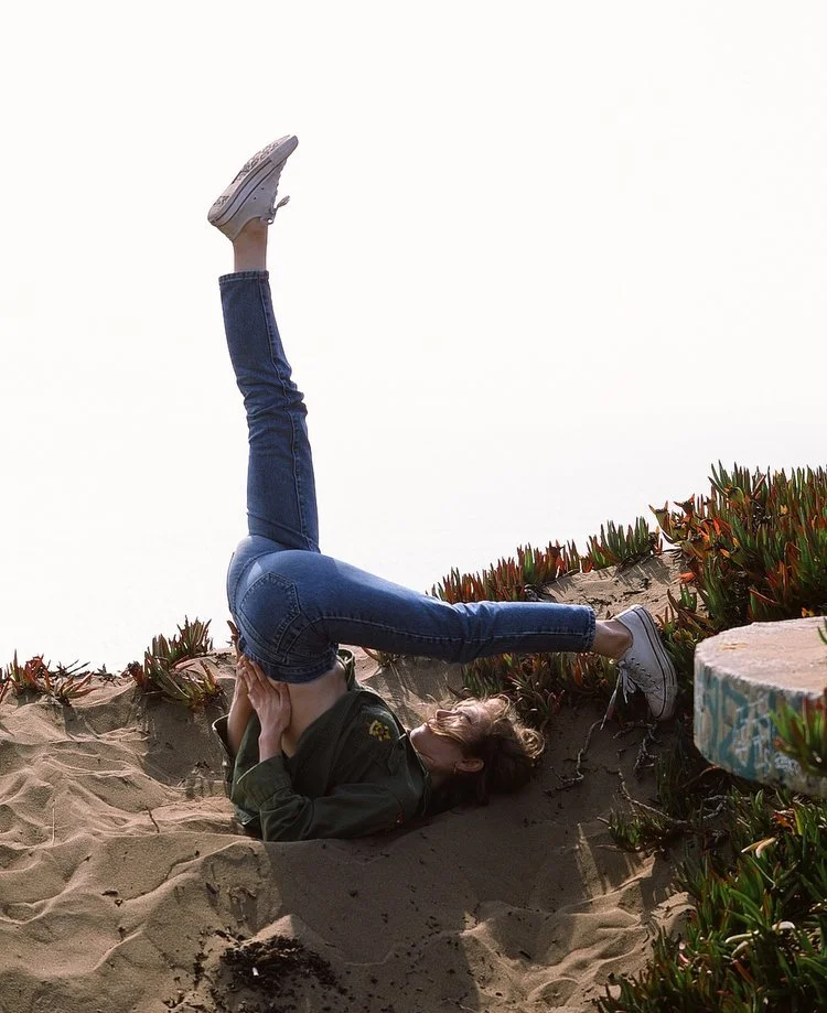 A person performing a headstand on a sandy beach, with their legs bent at the knees and feet pointing upwards, near some plants and graffiti.