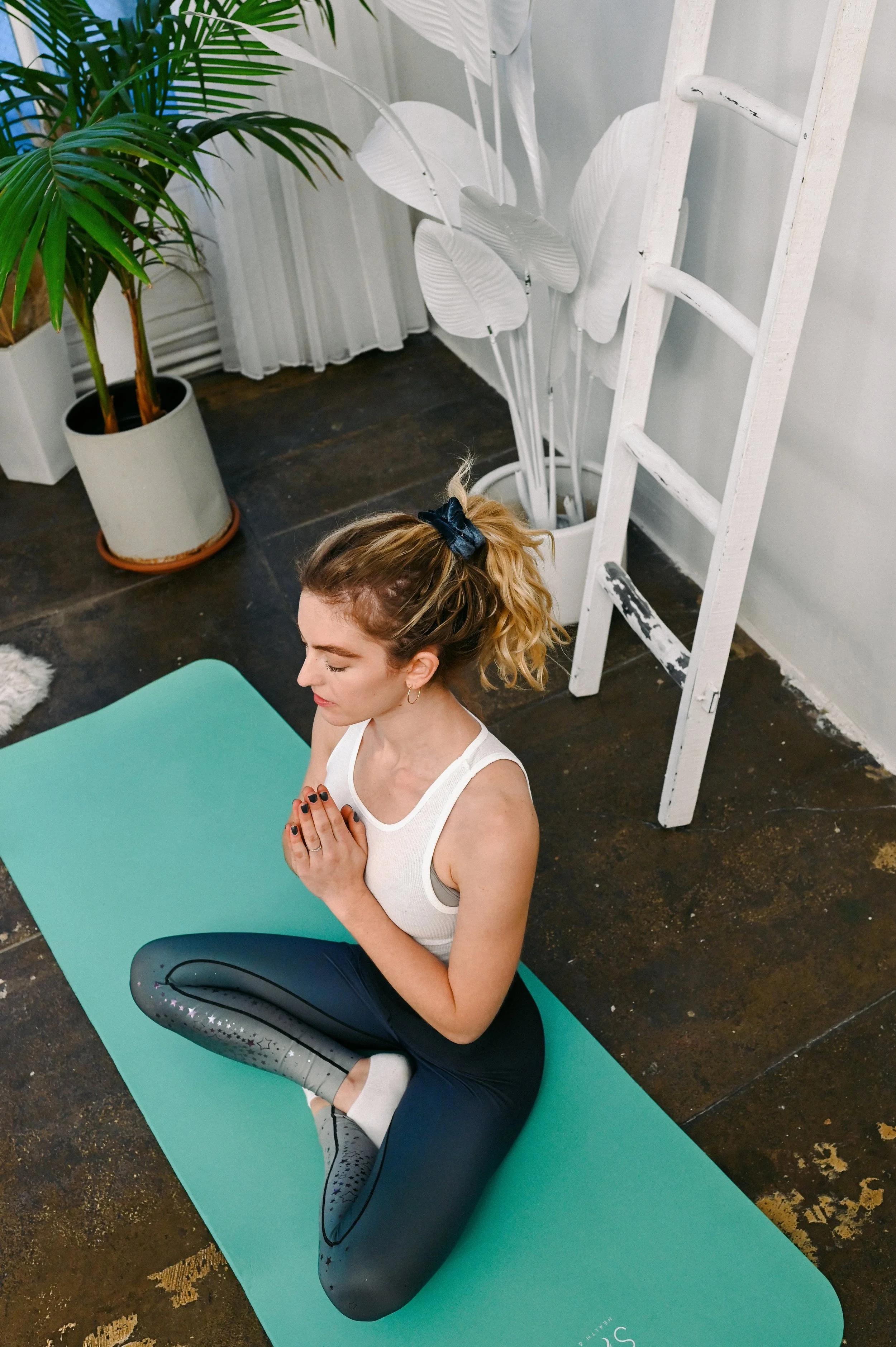 A woman practicing yoga indoors on a turquoise mat, sitting cross-legged with her hands in prayer position, eyes closed, surrounded by houseplants and a white ladder.
