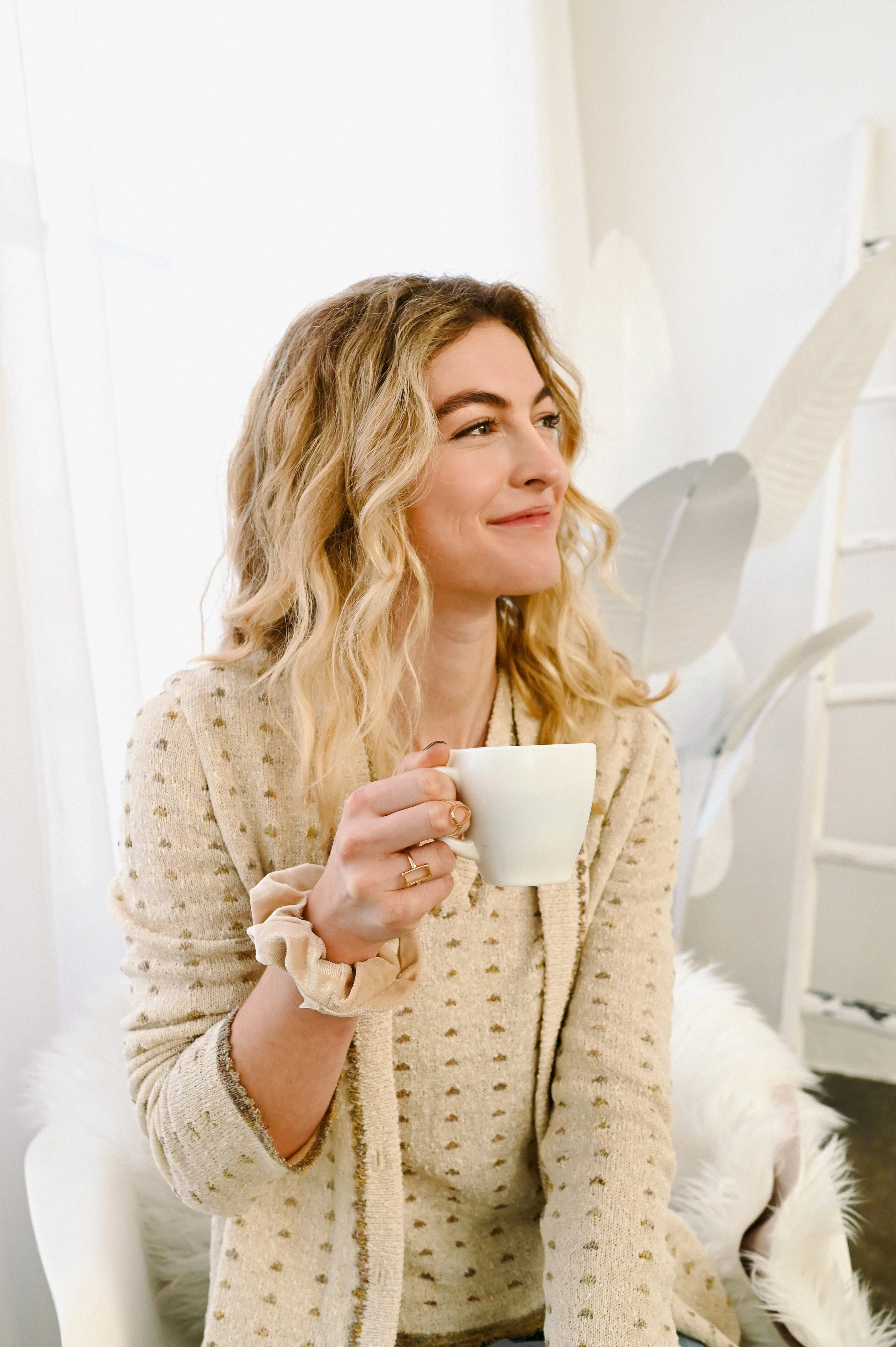 A woman with blonde, wavy hair is sitting on a chair, holding a white coffee mug, and smiling softly while looking to the side. She is wearing a beige, dotted cardigan, and is in a bright, cozy indoor setting with white walls and decorative leaves in