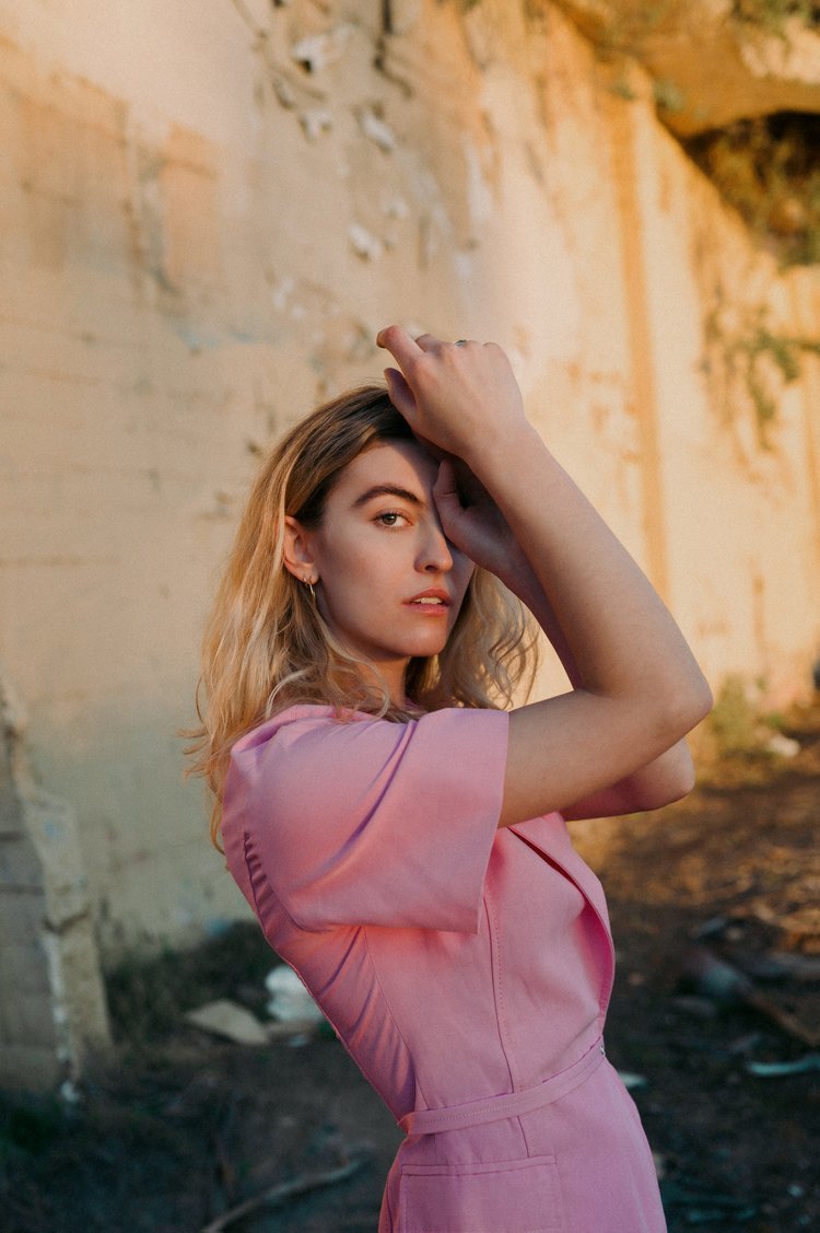 A woman with blonde hair and fair skin, wearing a pink dress, posing outdoors against a weathered brick wall during sunset.