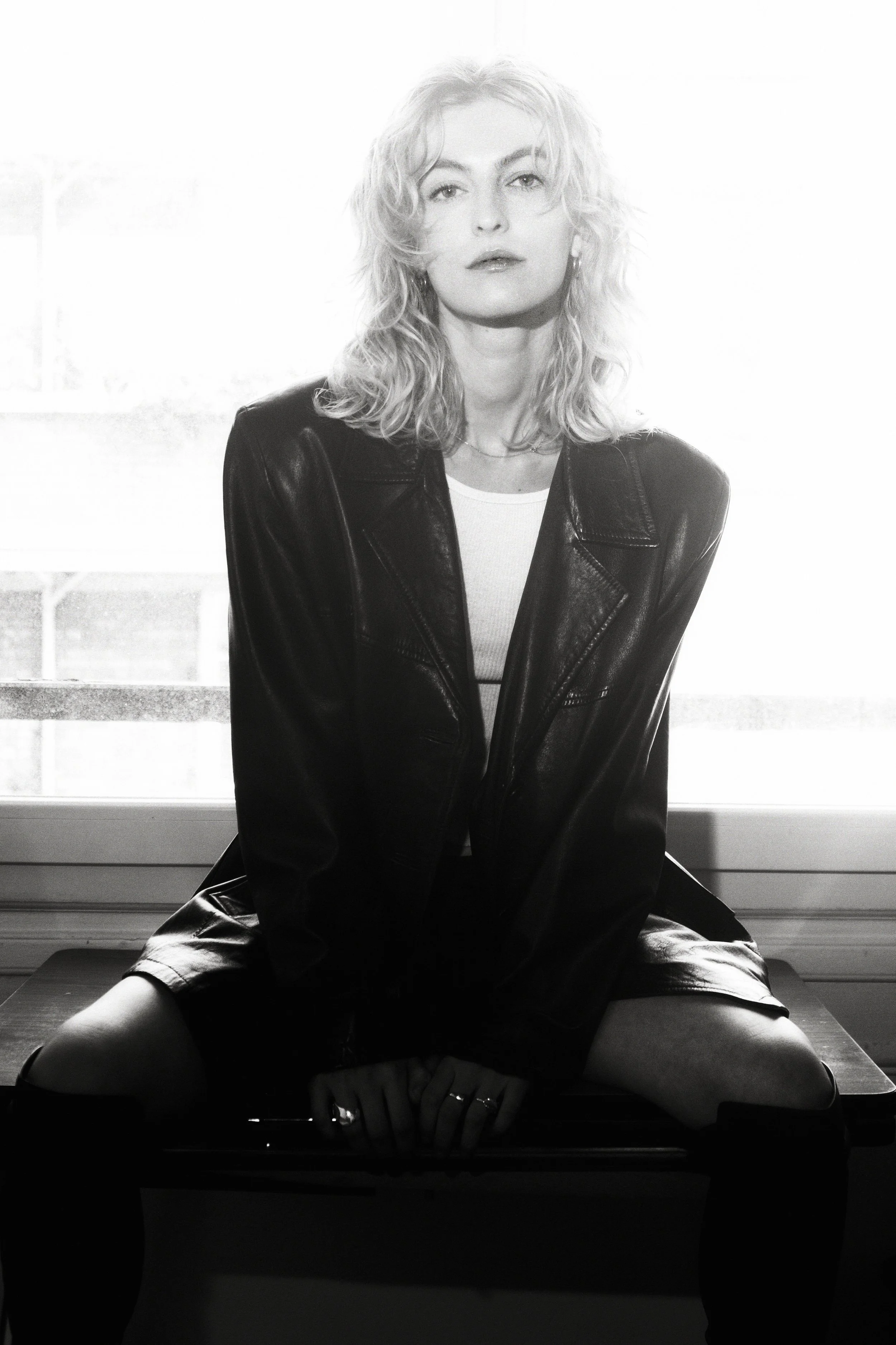 Black and white photo of a woman with curly hair, wearing a leather jacket, sitting on a table by a window.