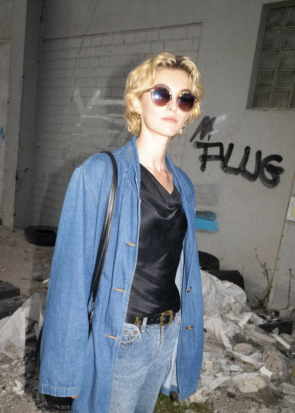 A young woman with curly blonde hair wearing sunglasses, a black top, a blue denim jacket, and jeans, standing outdoors in front of a graffiti-covered wall and debris.