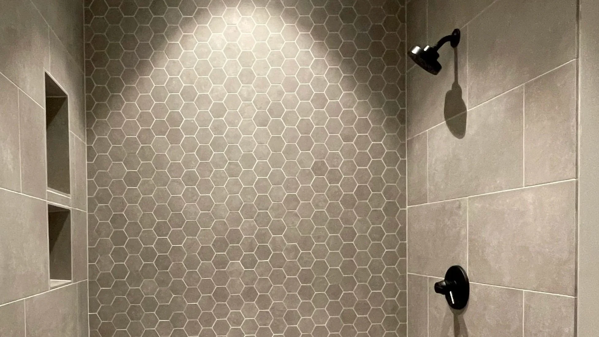 The shower area features hexagon tiles on the back wall and larger rectangular tiles on the side walls. There is a black showerhead on the upper right and a black handle or faucet below it. Built-in niche shelves are on the left side.