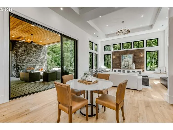 Open-concept living and dining area with large windows, modern furniture, and a view of an outdoor patio with seating and fireplace.