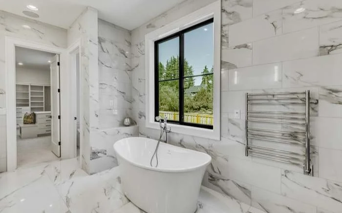 Modern bathroom with white marble walls and floor, a freestanding bathtub, a window showing an outdoor view, and a heated towel rail.