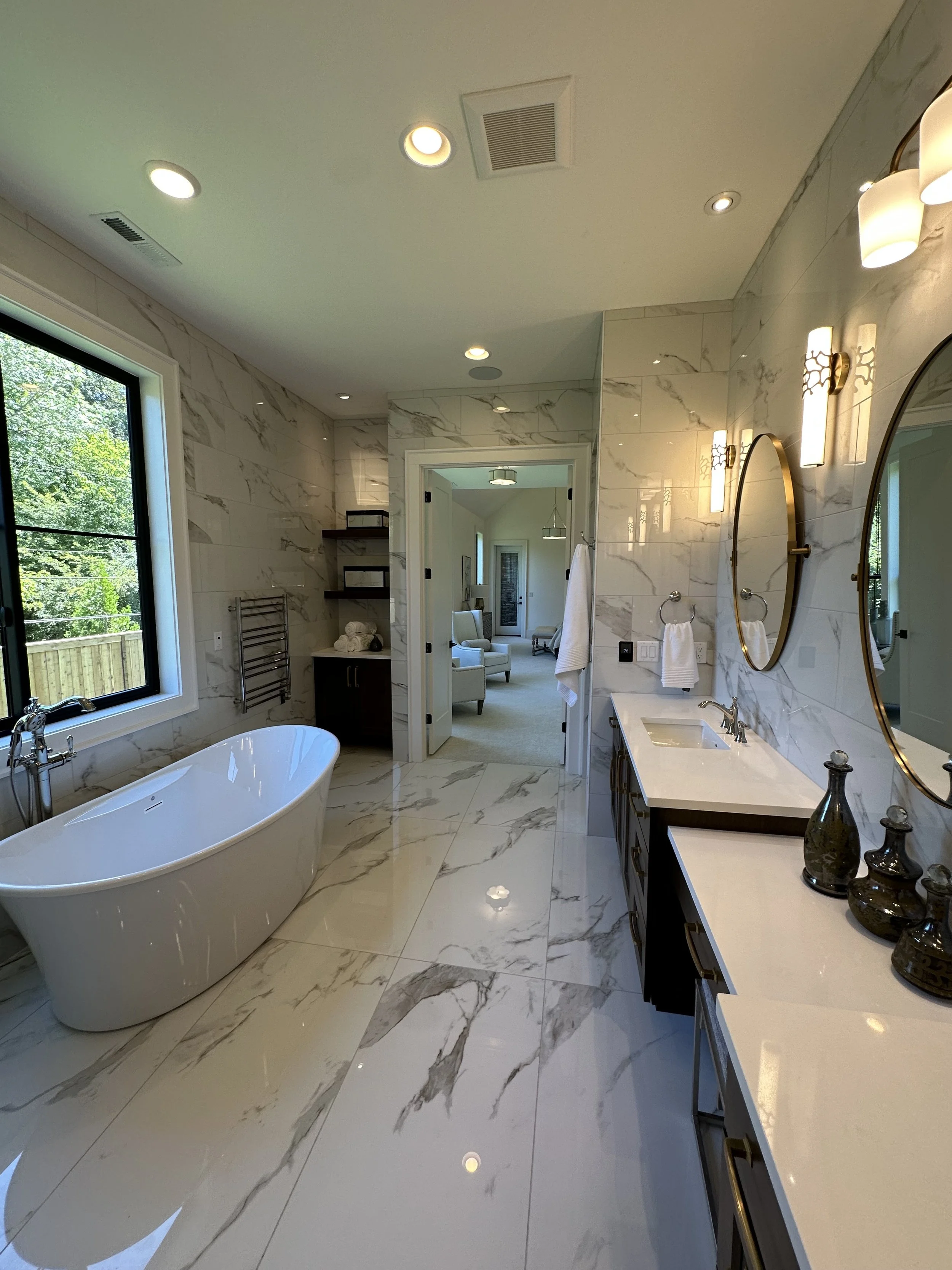 Luxury bathroom with marble flooring and walls, freestanding bathtub by a window, double vanity with two round mirrors and modern lighting fixtures, decor vases on the counter, and an adjoining room with seating visible through the doorway.