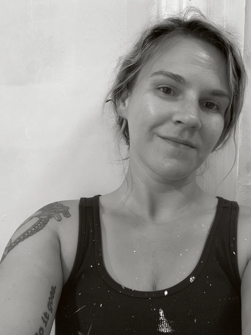 A woman with short hair, wearing a black tank top, taking a selfie against a plain wall, with some paint splatters on her shirt.