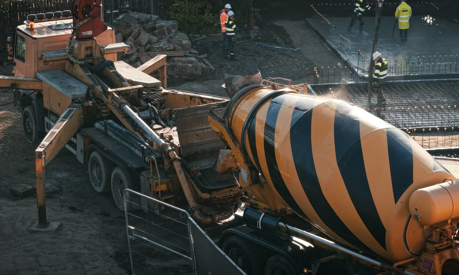 A construction site with a yellow and black striped concrete mixer truck and workers wearing safety gear and helmets pouring concrete.