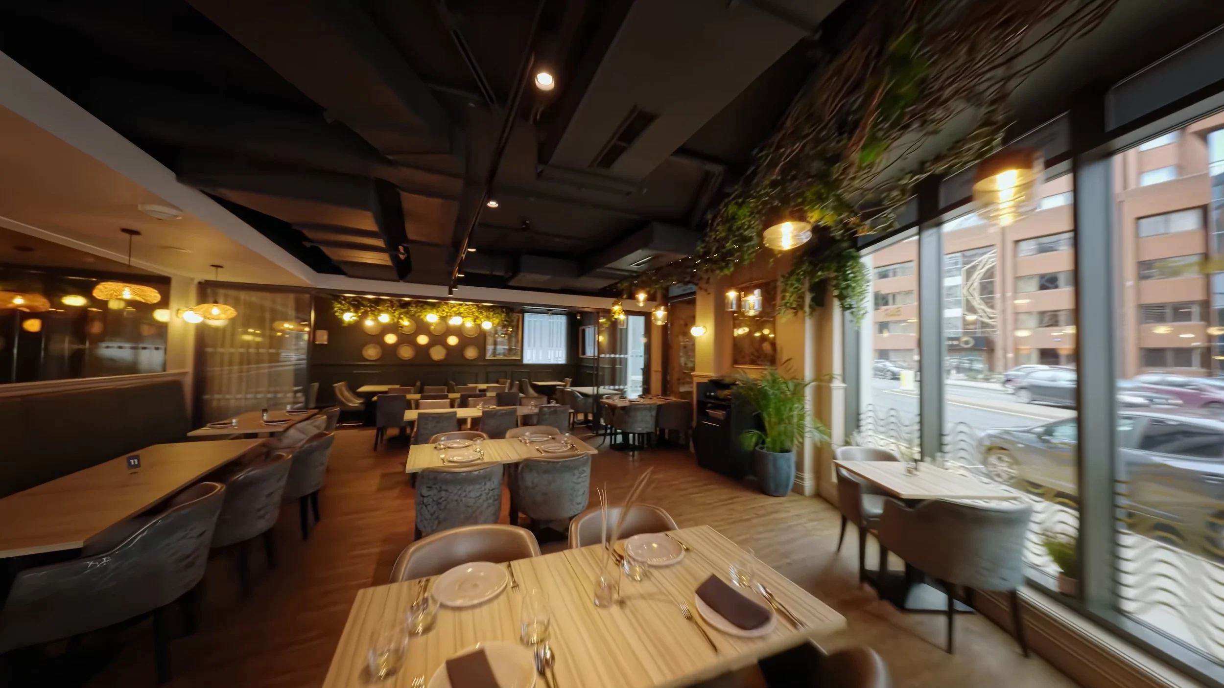 Elegant restaurant interior with large windows, modern black and gold lighting fixtures, a wine refrigerator, and neatly set tables with glassware and napkins.