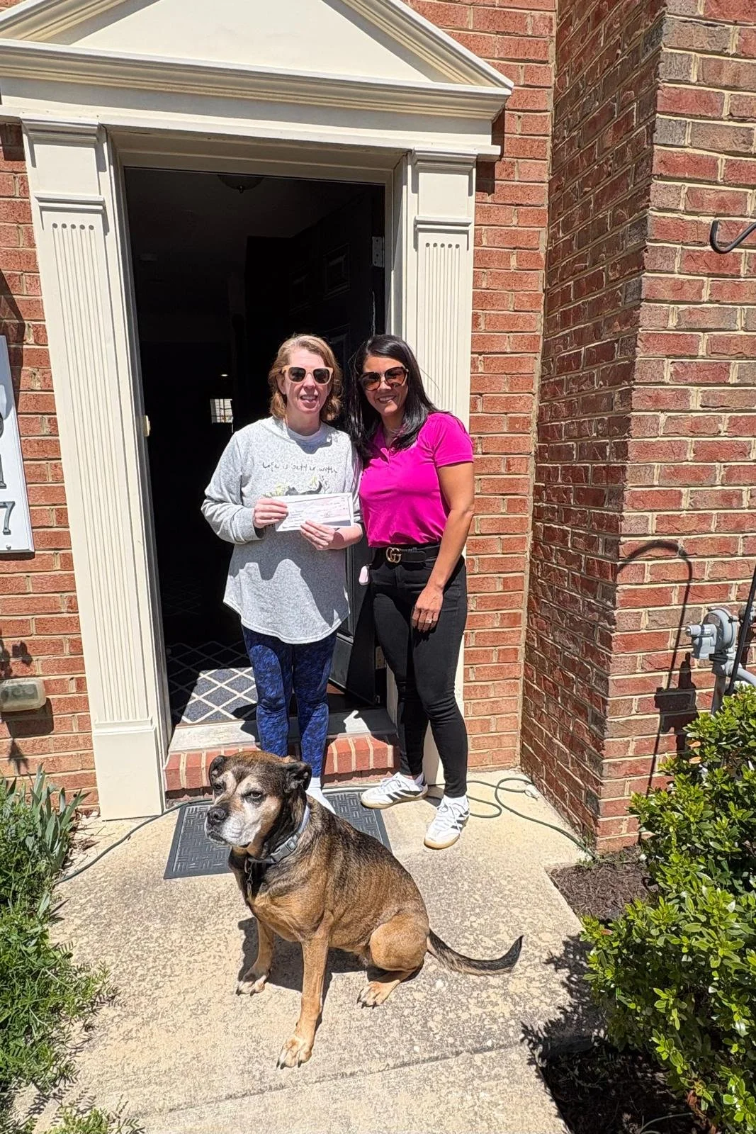 Two women and a dog. The homeowner in the grey  sweatshirt is holding a check given from Pink Roofing as a winner for a financial grant for the month of May. It's a warm sunny day and this is outside of her home. 