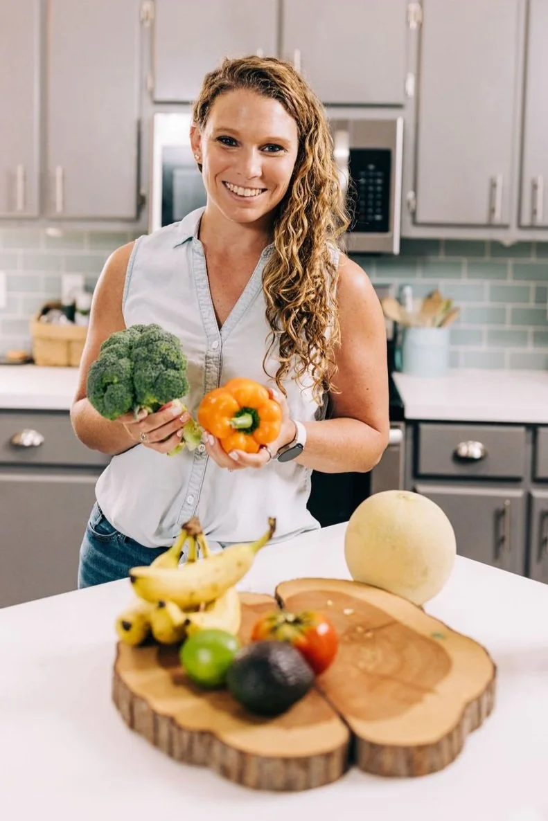 A woman smiling in a kitchen, holding a head of broccoli and a yellow bell pepper, with bananas, an avocado, a green apple, and a tomato on a wooden board on the counter in front of her.