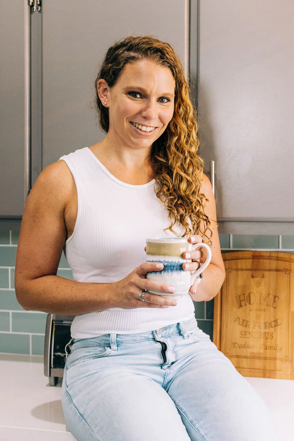 A woman with long curly hair smiling while sitting on a kitchen counter, holding a colorful mug.