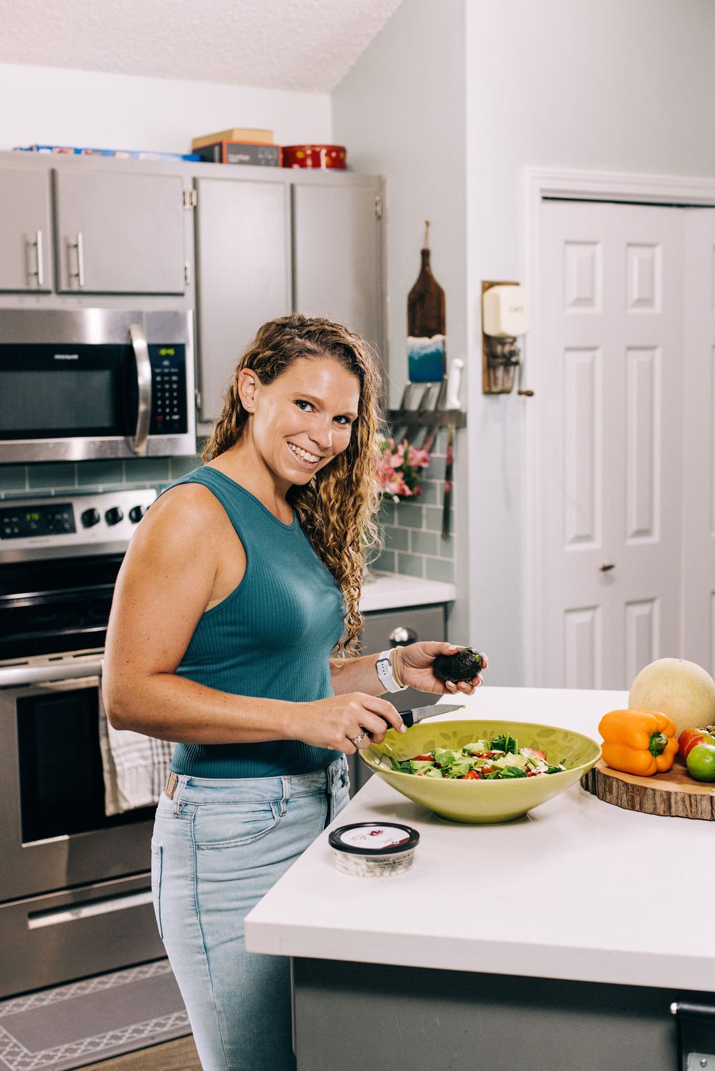 A woman with curly hair smiling while chopping an avocado in a kitchen, surrounded by vegetables and kitchen appliances.