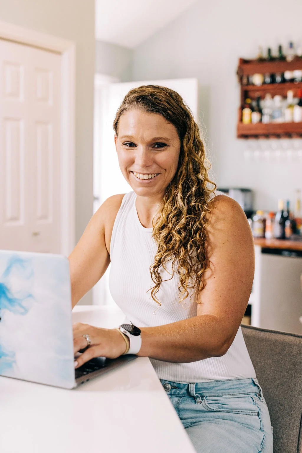 A woman with long curly hair smiling while working on a laptop in a kitchen.