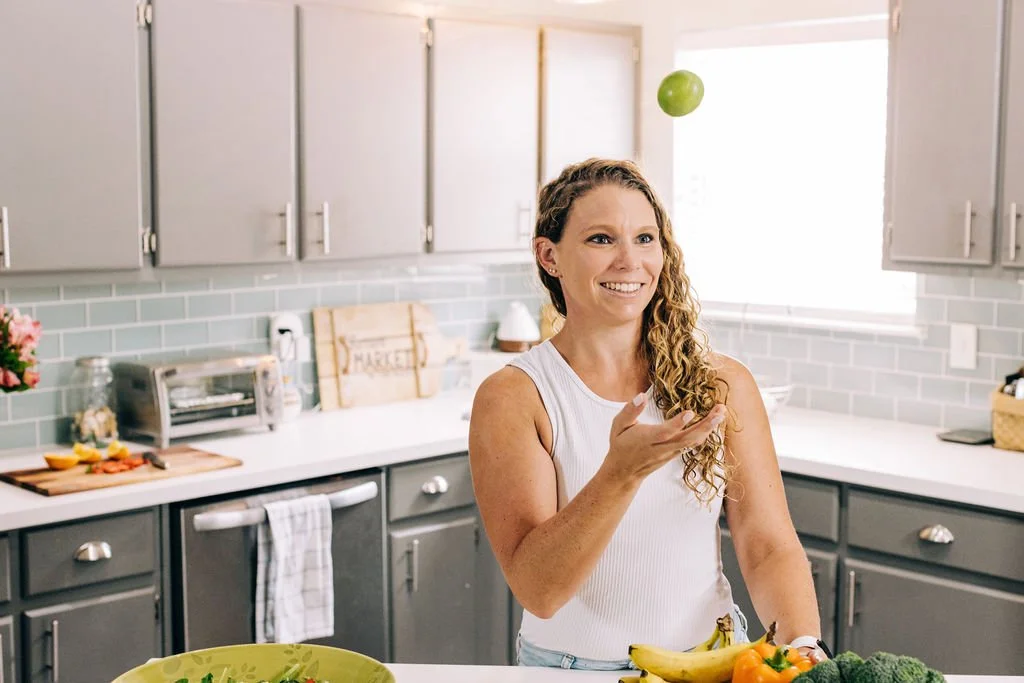A woman with long curly hair is in a kitchen, tossing a lime into the air with a smile, surrounded by a plate of bananas, broccoli, and peppers.