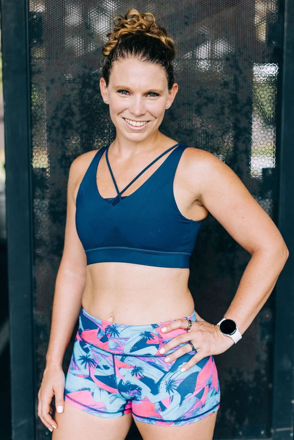 A smiling woman wearing a navy sports bra and colorful tropical print shorts, standing with one hand on her hip in front of a dark perforated metal background outside.