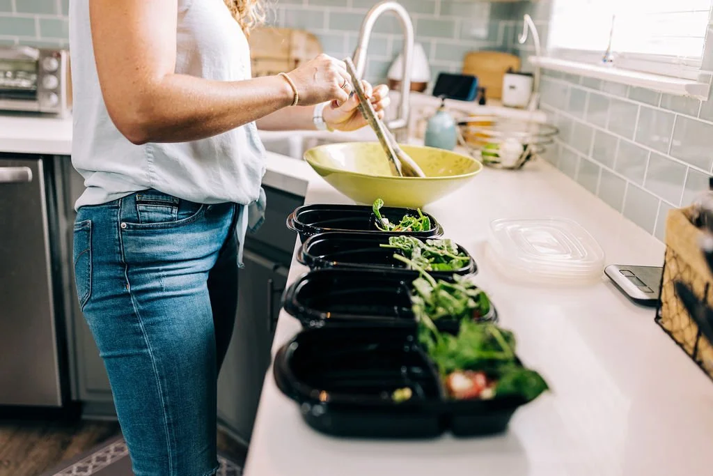 A woman meal prepping in a kitchen, filling black containers with leafy greens, with a large green mixing bowl nearby.