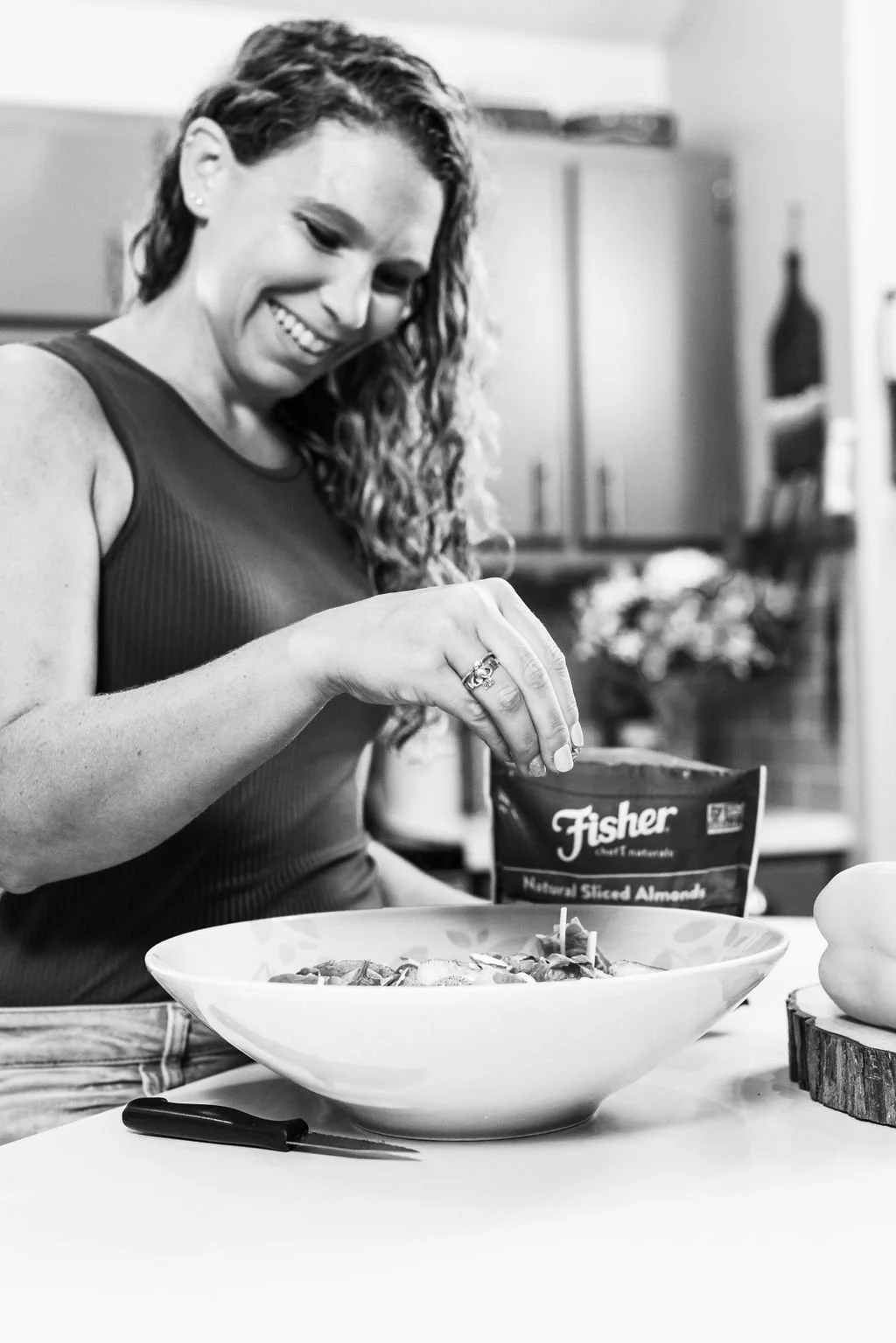 A woman with curly hair smiling while preparing a salad in a kitchen, pouring almonds from a packet into a large bowl.