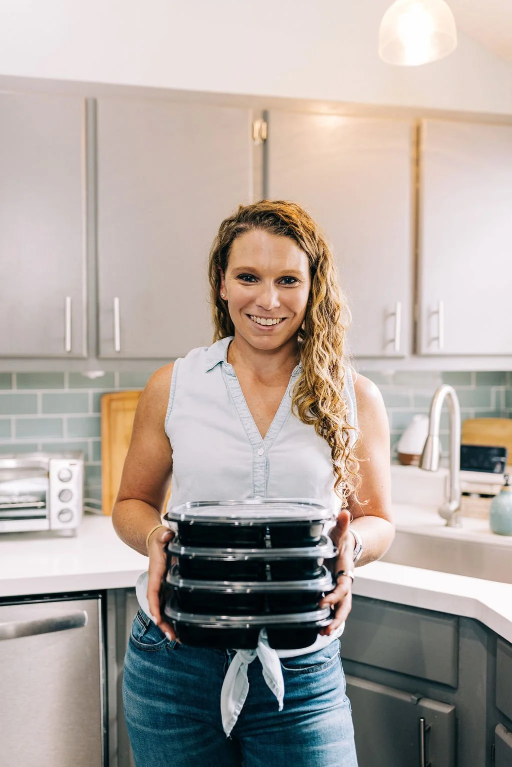 A woman with long curly hair smiling and holding a stack of four black takeout containers in a modern kitchen.