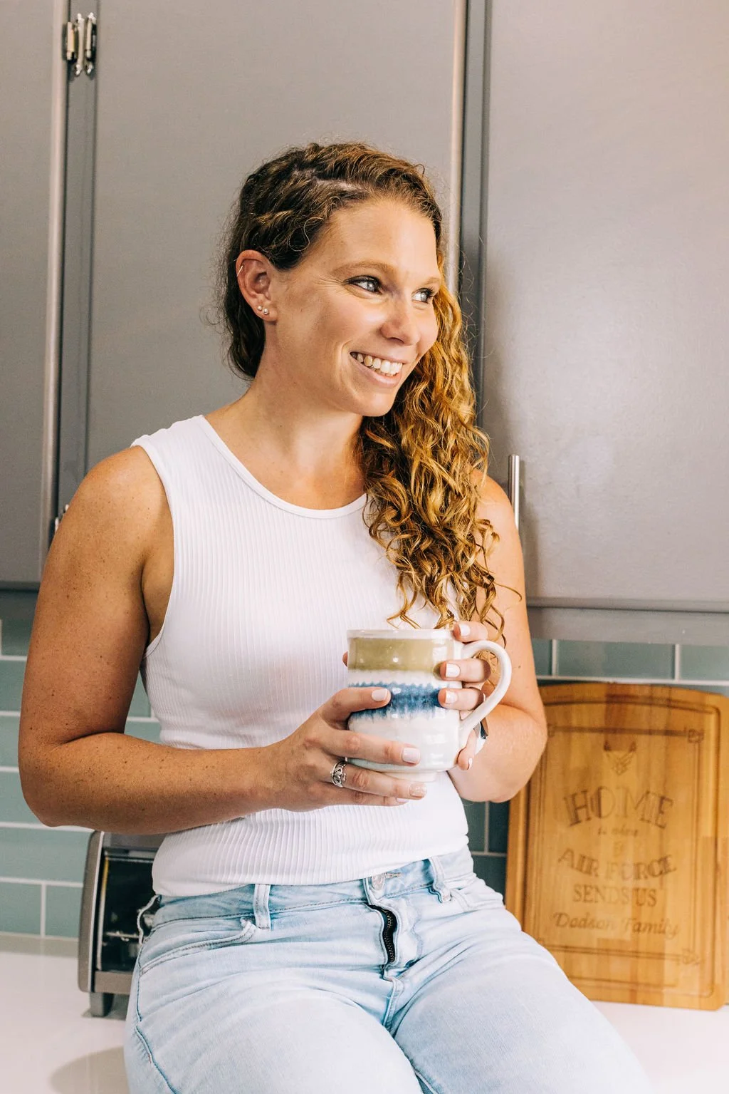 A woman with blonde, curly hair smiling and holding a mug, sitting on a kitchen counter.