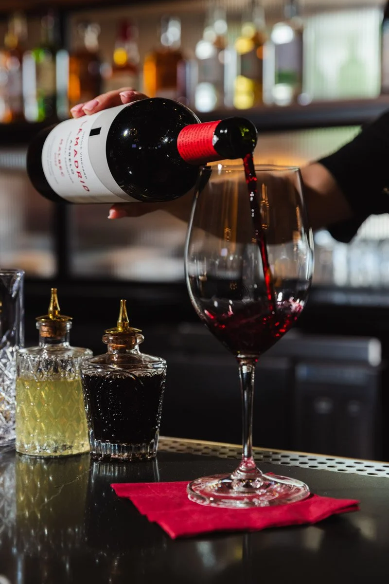 A bartender pours red wine into a wine glass on a bar counter with various bottles and condiments in the background.