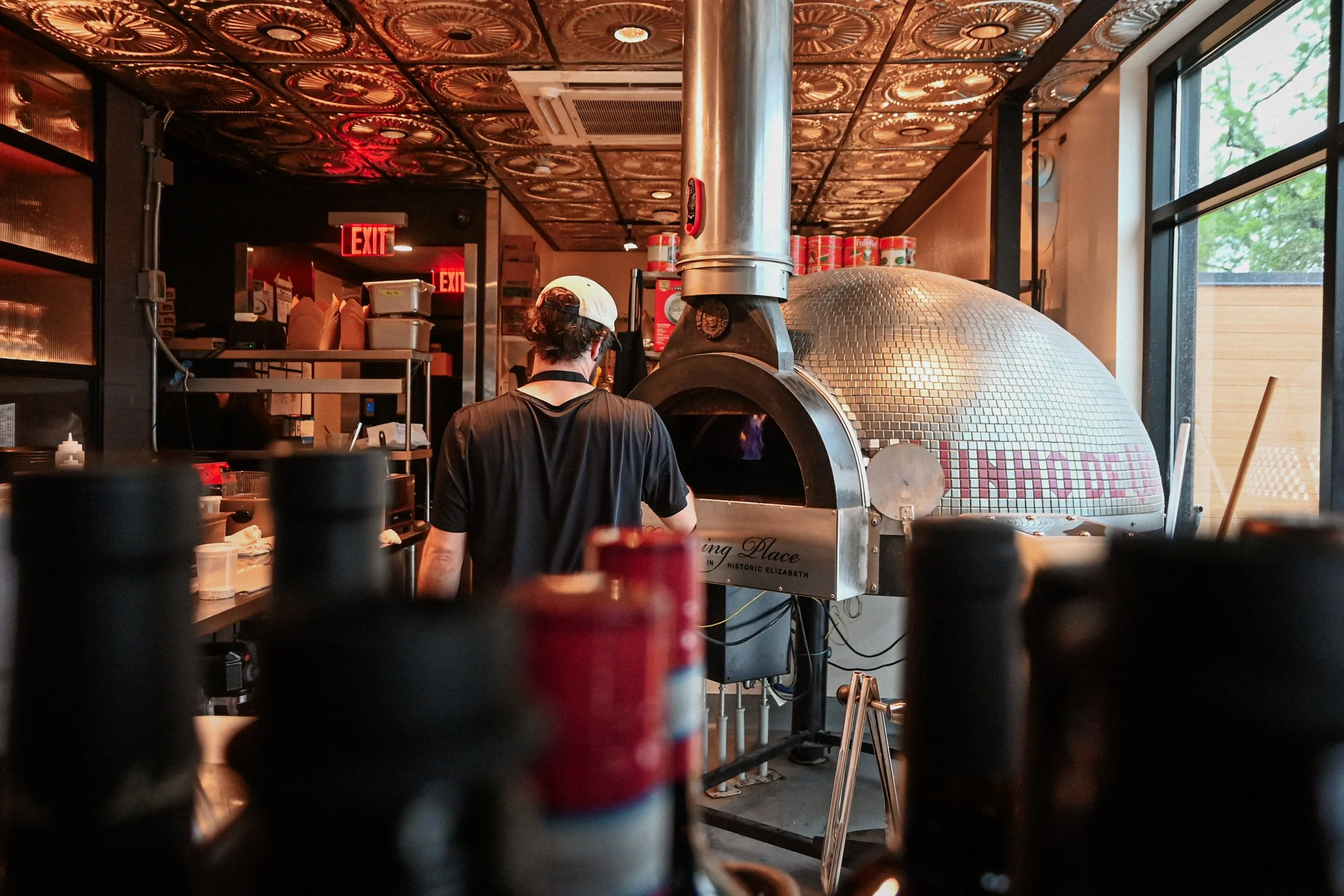 A person with curly hair wearing a white cap and dark shirt working in a pizzeria kitchen, with a wood-fired oven in the background. The kitchen has a decorative copper ceiling and large windows letting in natural light.