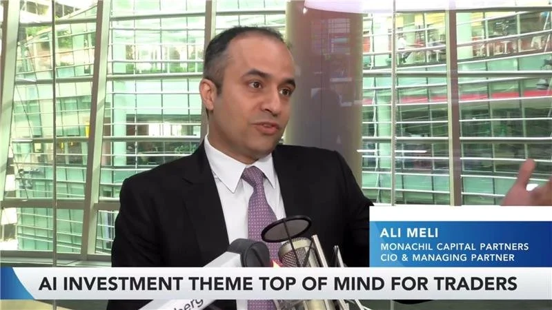 Ali Meli, CIO & Managing Partner at Monachil Capital Partners, speaking at a press event with multiple microphones in front of him, indoors with large glass windows showing a reflection of nearby buildings.