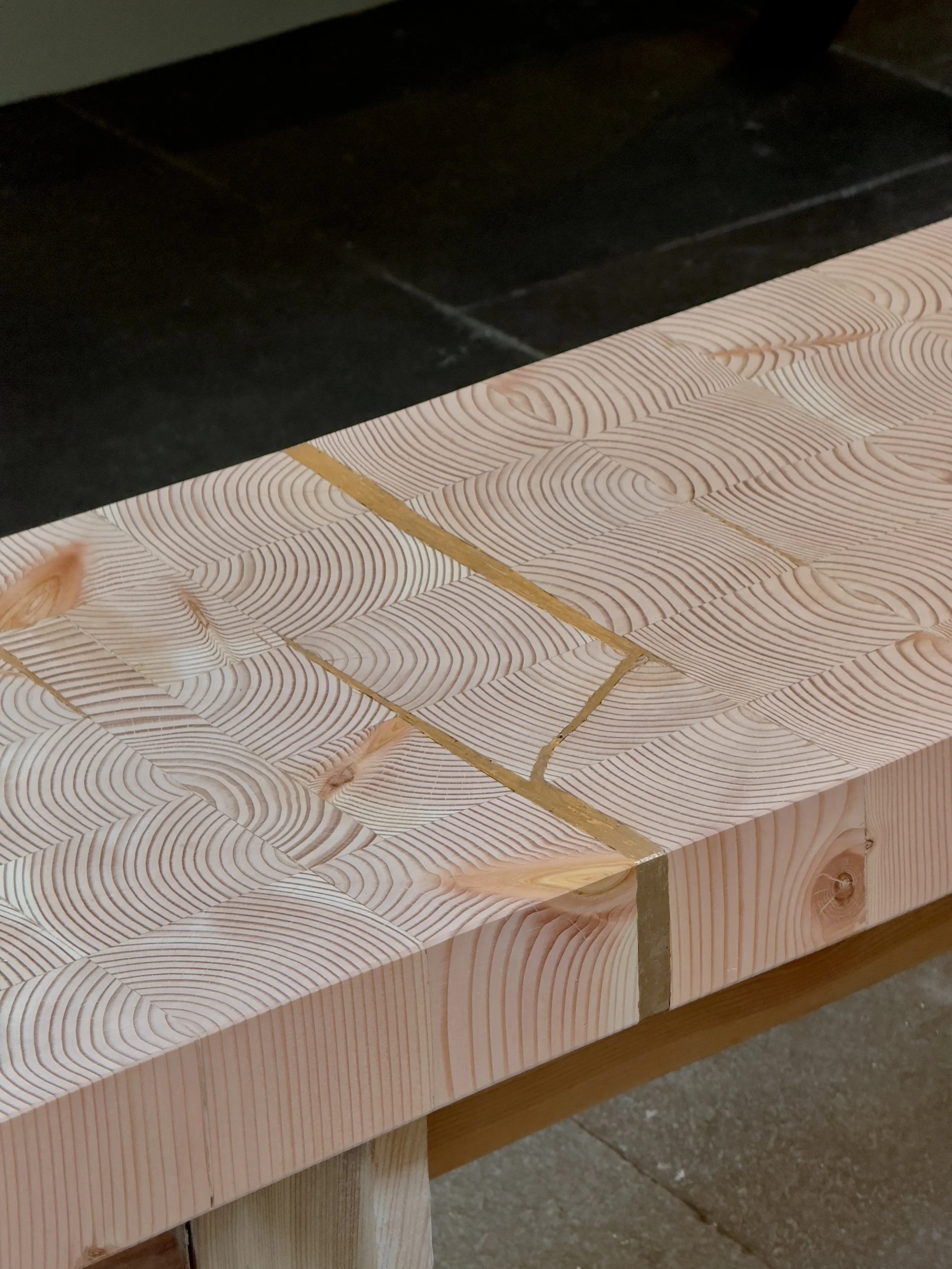 Close-up of a wooden table with a patterned, textured surface and visible wood grain, set against a dark tiled floor. Kintsugi bench.