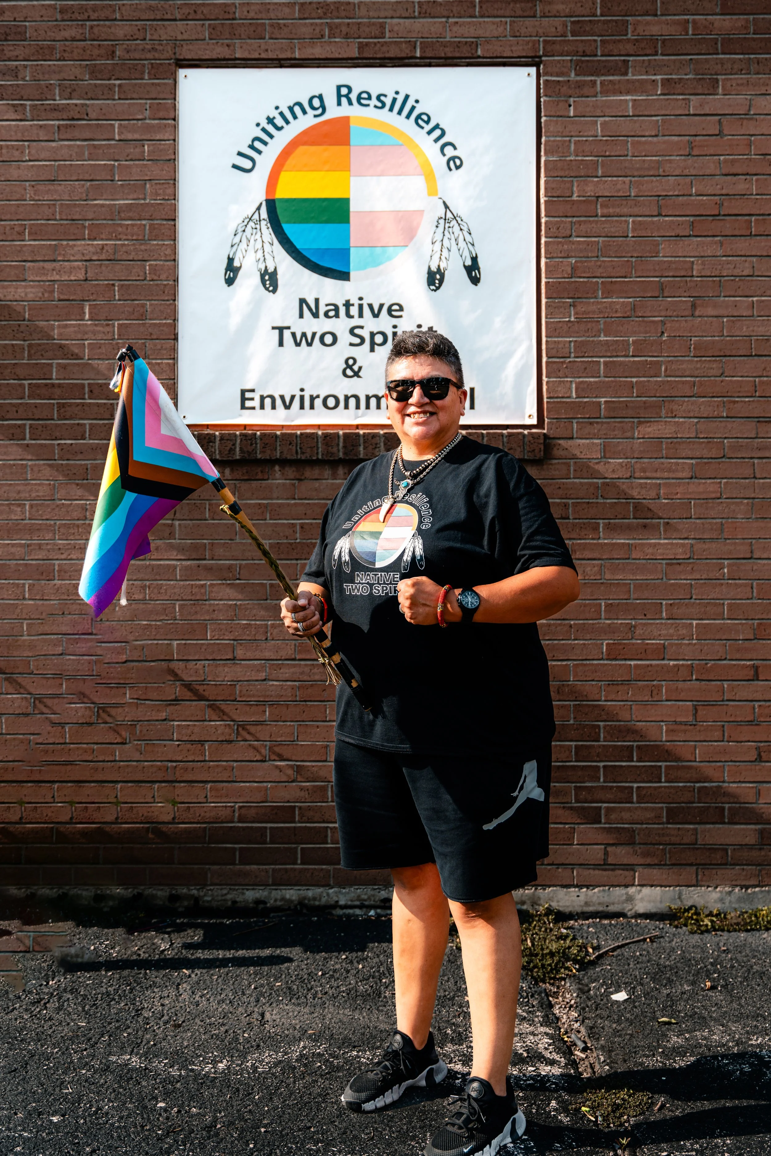 Muffie stands with a pride flag in front of a sign with a Two-Spirit medicine wheel and text "Uniting Resilience".