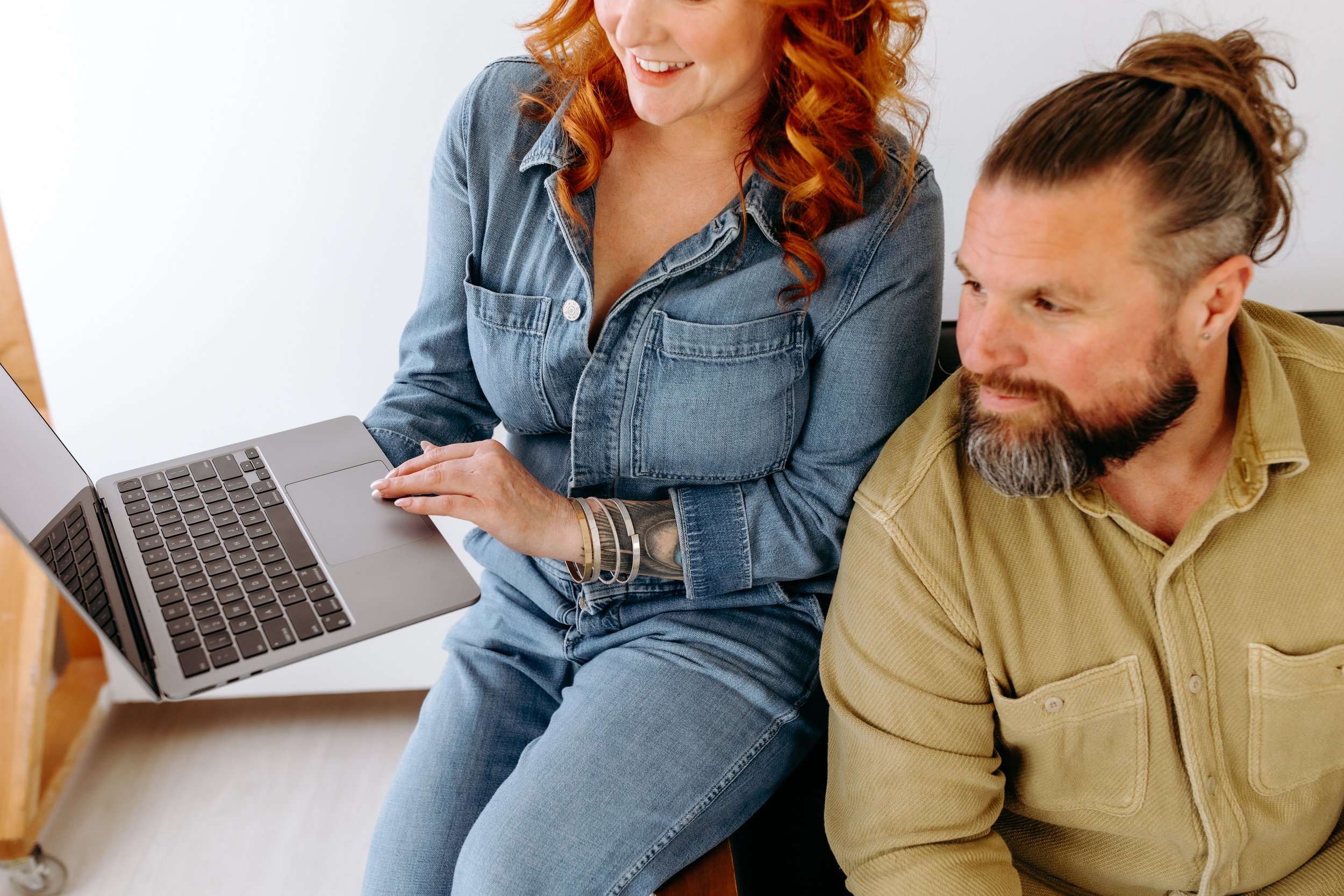 A woman with curly red hair and a man with a beard and long hair in a top knot working together on a laptop.