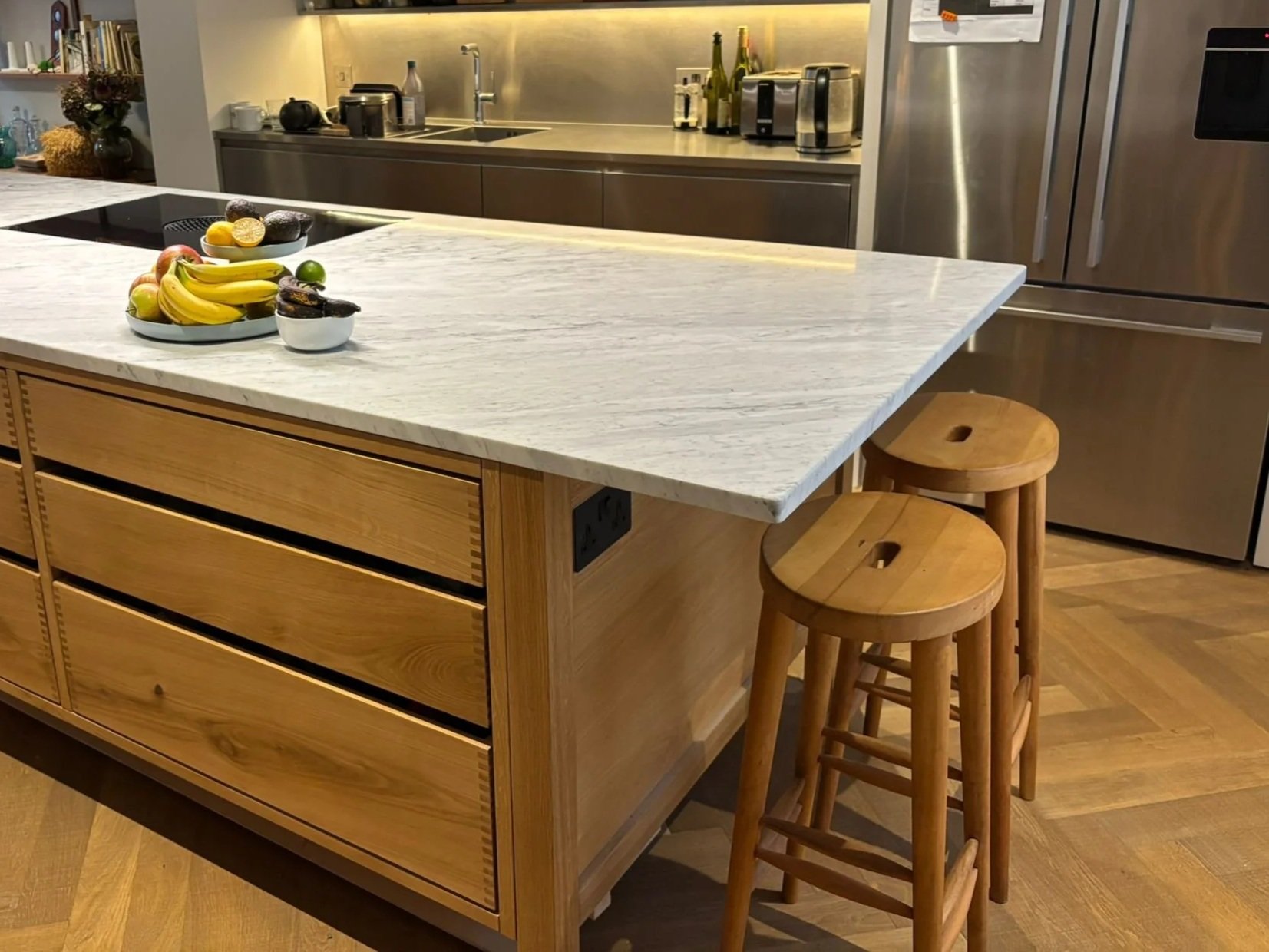 A modern kitchen with a marble countertop island, two wooden stools, stainless steel appliances, and a bowl of fruit on the island.