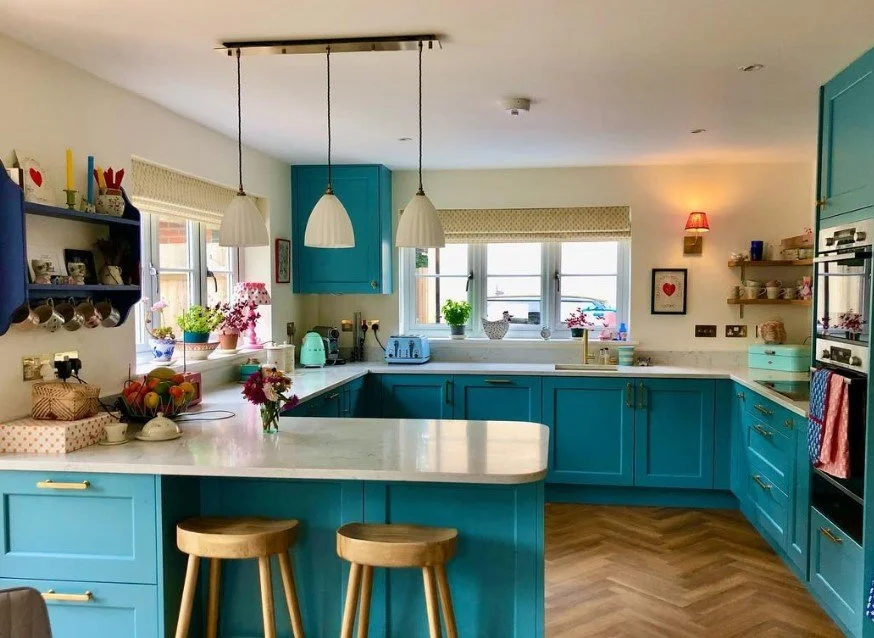 Bright kitchen with blue cabinets, white countertops, wooden bar stools, and various potted plants, flowers, and kitchen items on the counters and shelves.