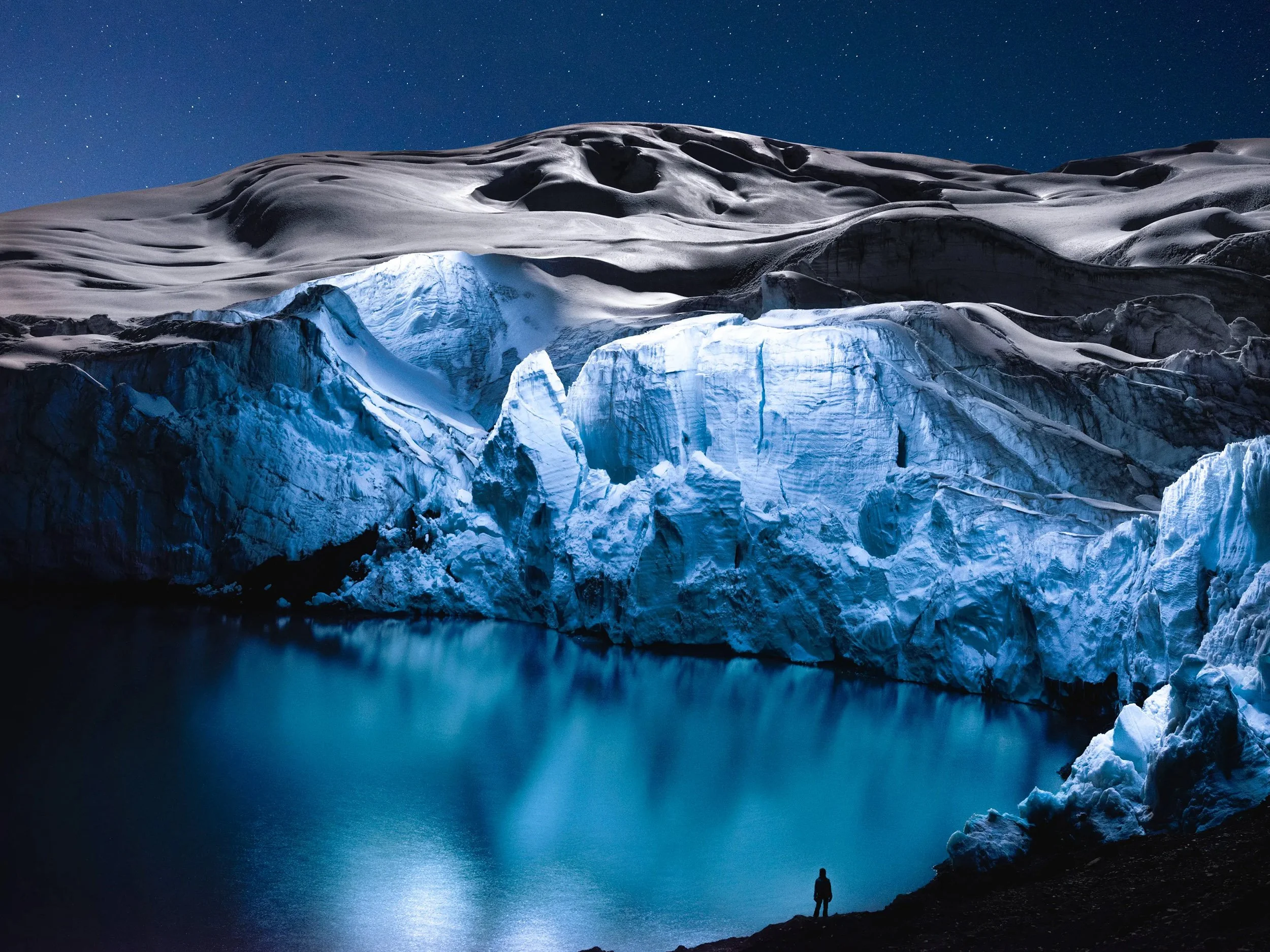 A lone figure stands in the night, viewing a lit glacier - in one of Reuben Wu's surreal photographs.