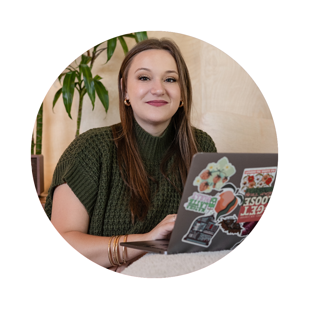A young woman with light skin and long brown hair sitting at a table with a laptop decorated with various stickers, smiling at the camera. She is wearing a green knit sweater and gold jewelry. There is a plant in the background.