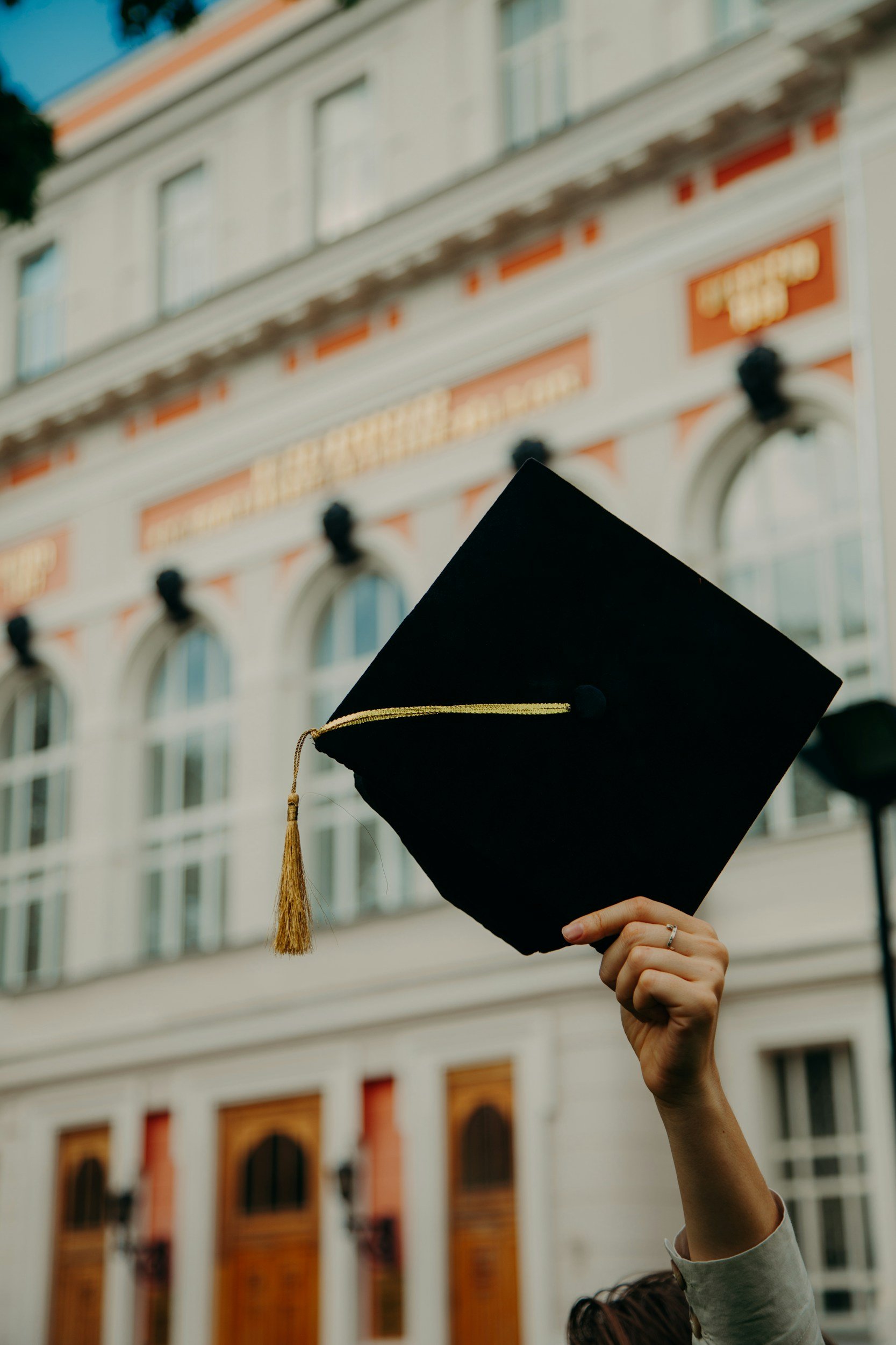 Person holding a black graduation cap with a gold tassel in front of a historic building with arched windows and decorative architecture.