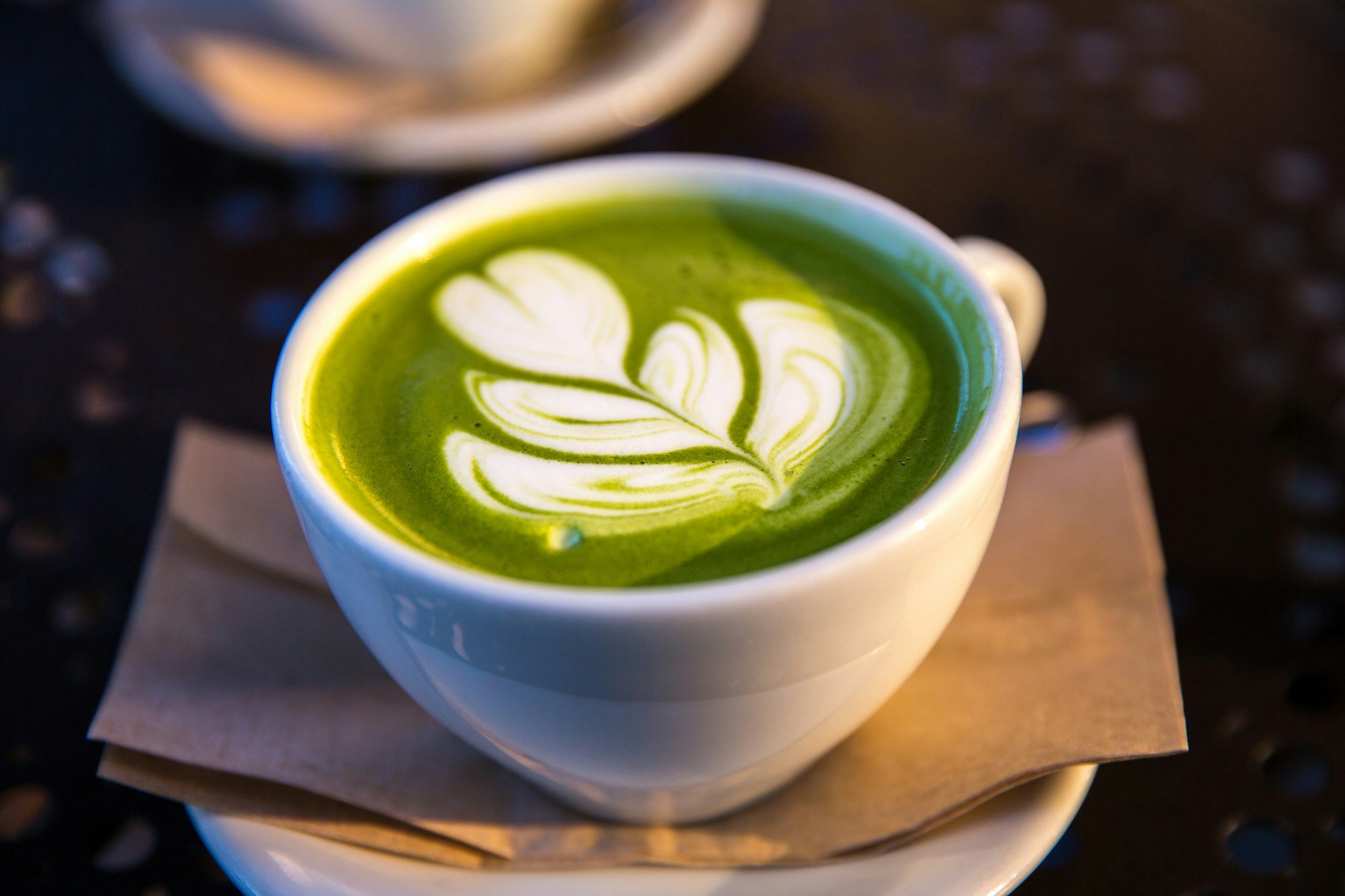 A cup of matcha green tea latte with latte art in a white ceramic cup on a saucer, on a napkin, with a blurred background.