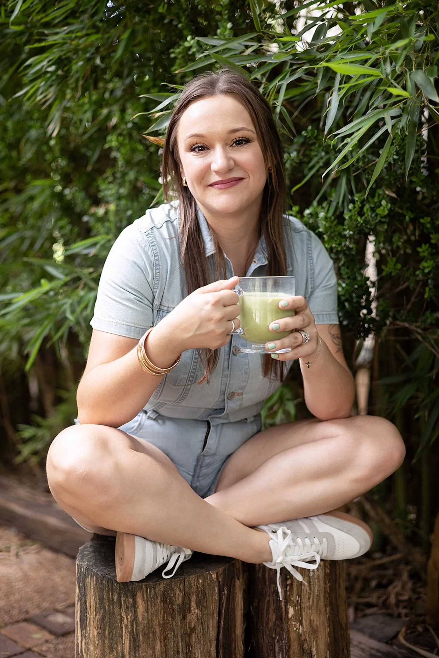 Fractional CMO Morgan sits criss cross on a wooden log. She's wearing a denim romper and holding a glass of matcha tea while smiling warmly at the camera.
