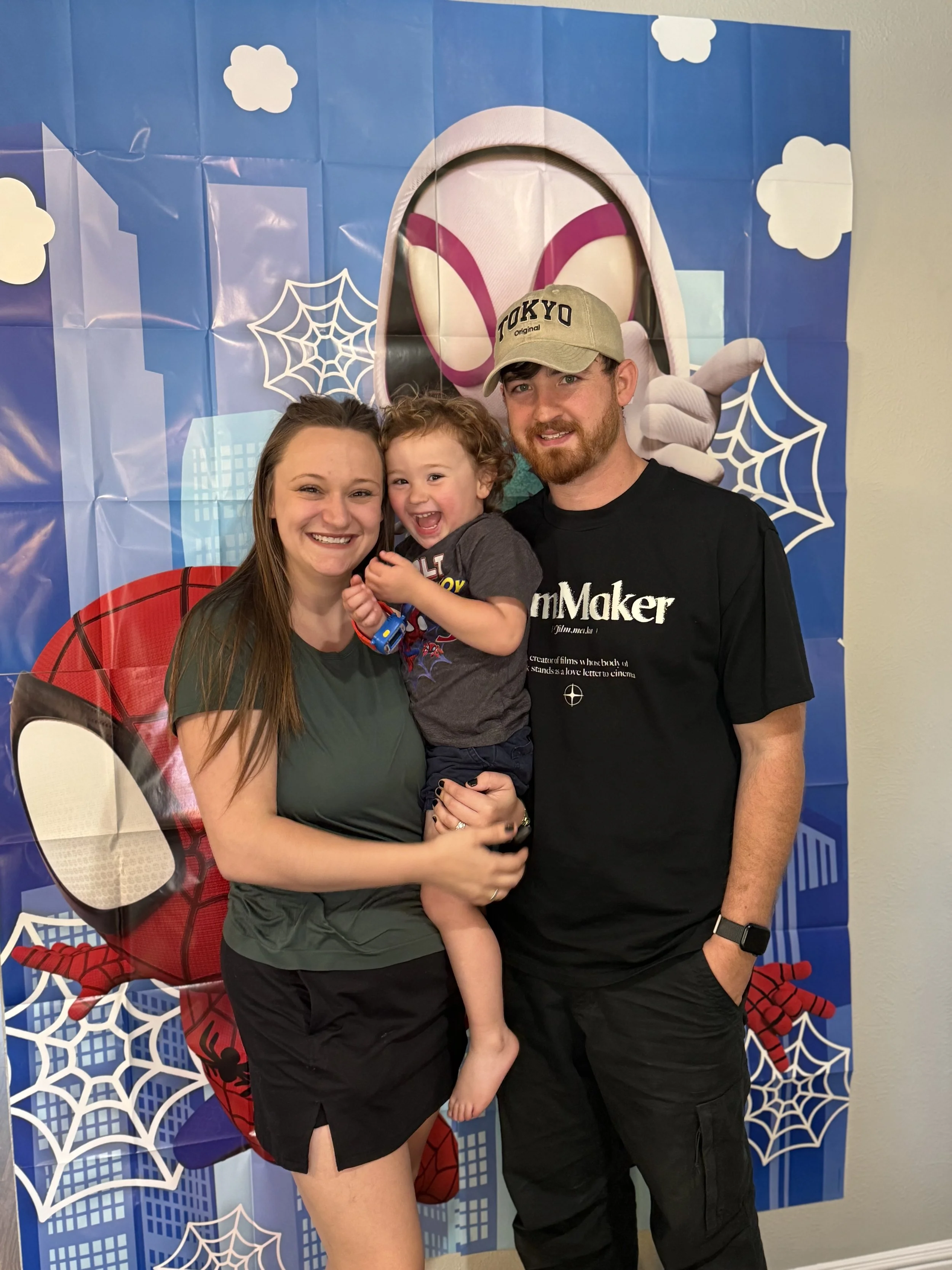 Family of three posing in front of a Spider-Man and Spider-Man-themed backdrop with graphics of clouds, webs, and city buildings. The woman is holding the young boy who is smiling, and the man is standing next to them. The boy wears a gray T-shirt, and the woman and man wear dark clothing.