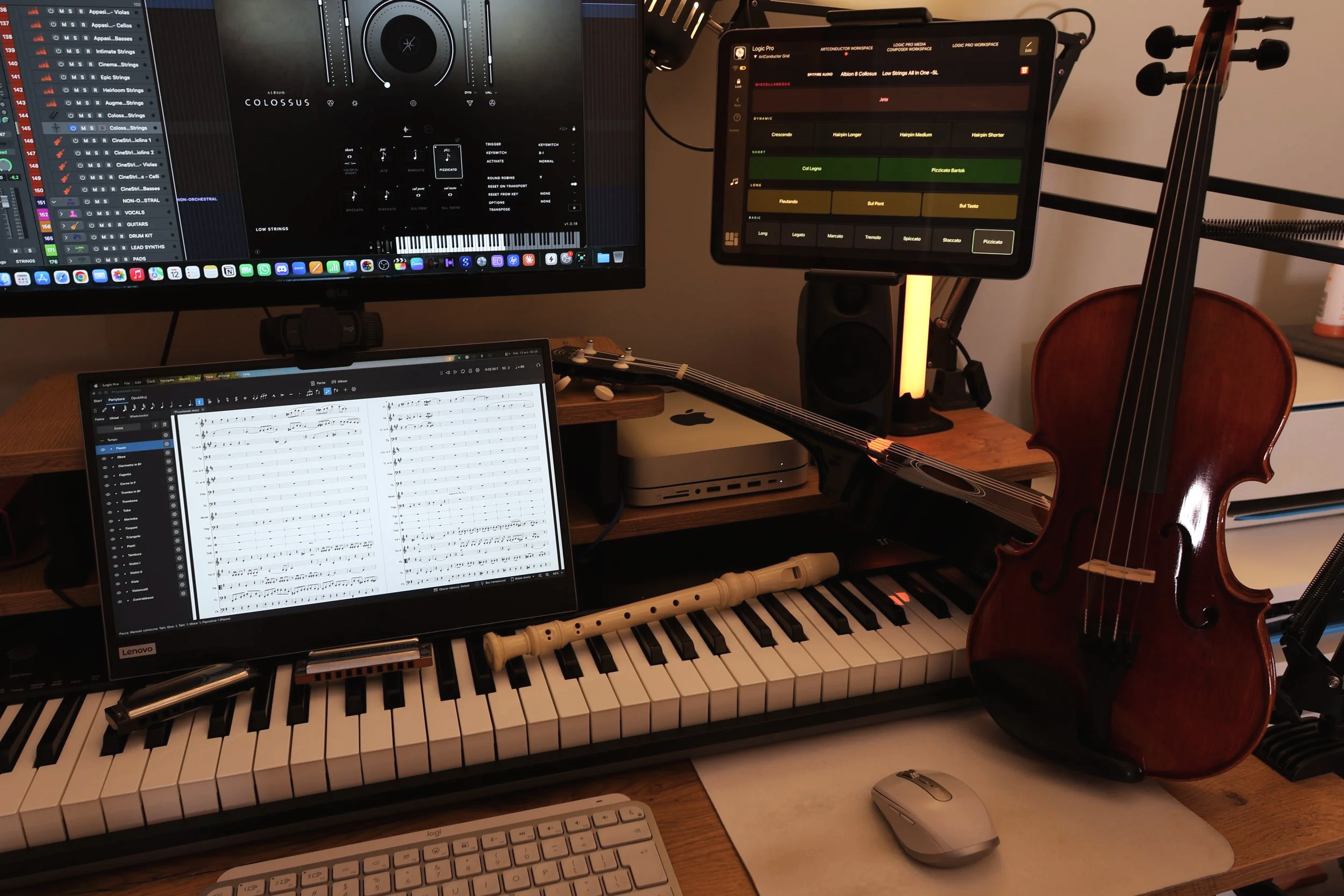 The photo shows Grzegorz Drozdowski's home studio desk with his Mac and music setup, with opened sessions in Logic Pro, Musescore Studio and Metagrid Pro. It also shows a few acoustic instruments the composer can play on a basic level, like violin