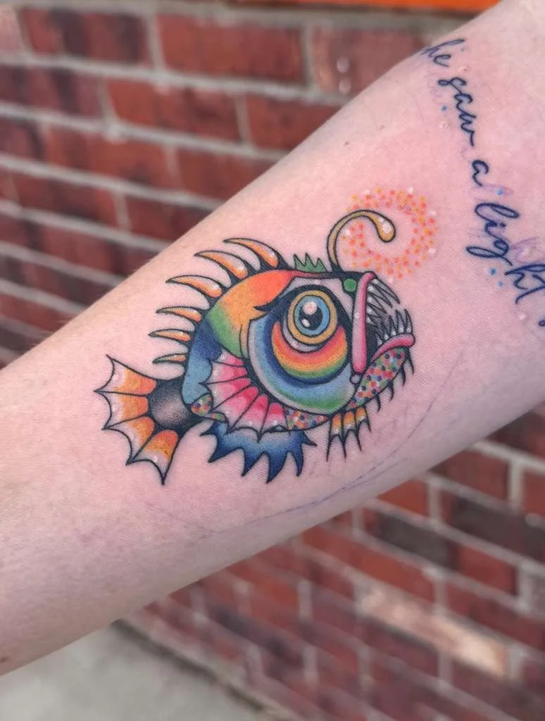 Colorful tattoo of a fish with rainbow colors and an exaggerated head, set against a brick wall background.